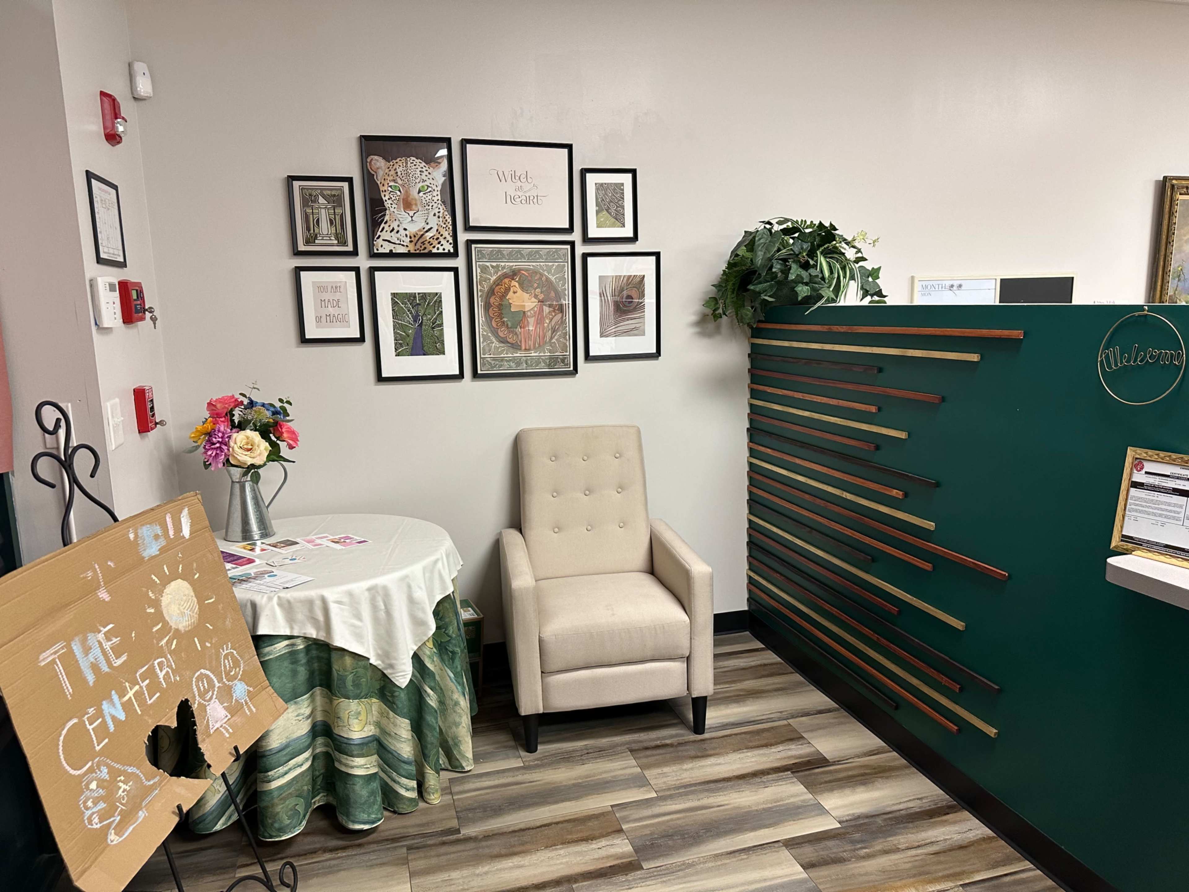 A small reception area with a chair, a table covered with a cloth and flowers, framed artwork on the wall, and a green reception desk with decorative strips.