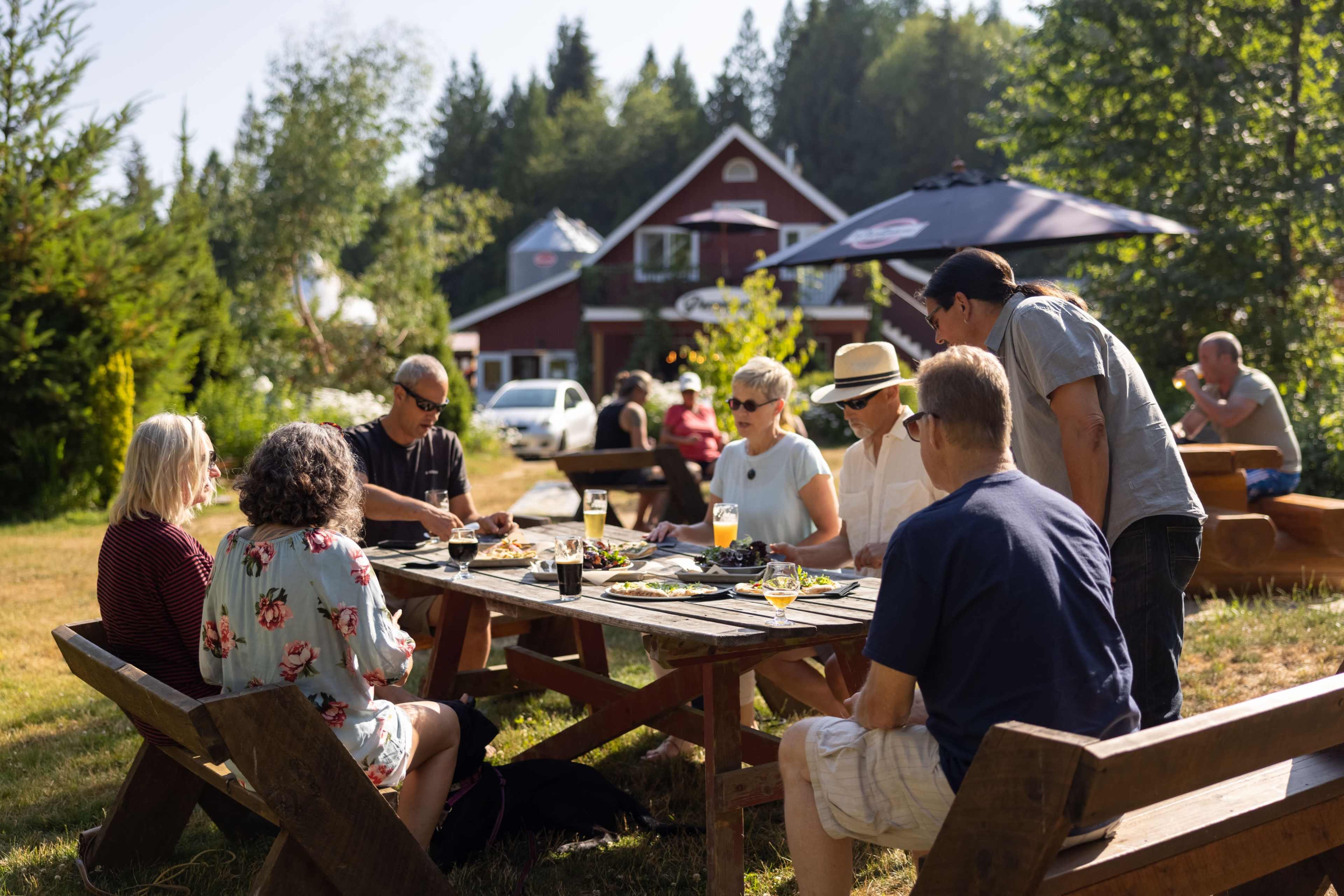 A group of people sits around a wooden table enjoying a meal outdoors at a scenic location with a red house in the background.