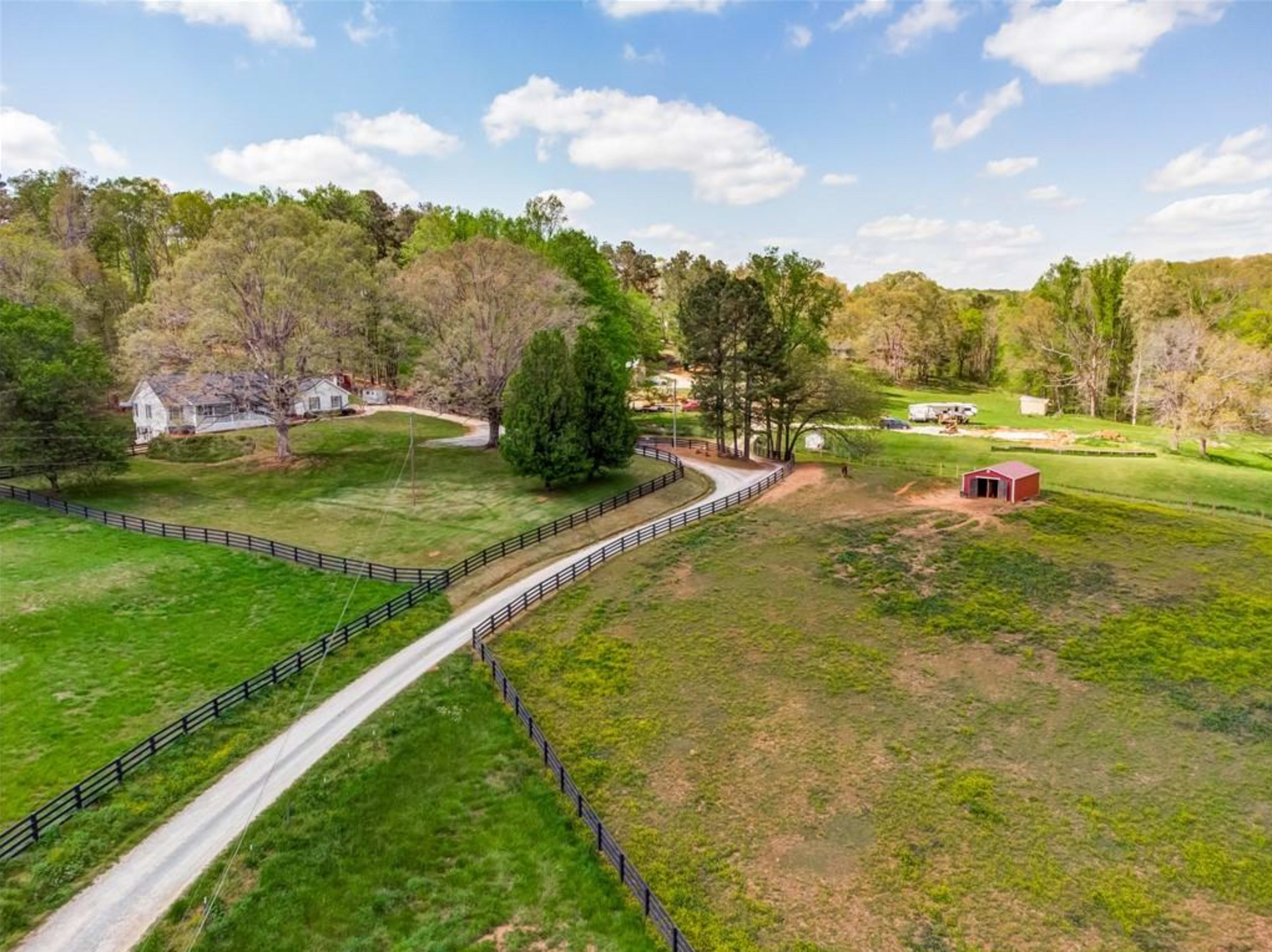 A rural landscape featuring a winding driveway bordered by green fields and trees, leading to a house and a red barn in the distance.