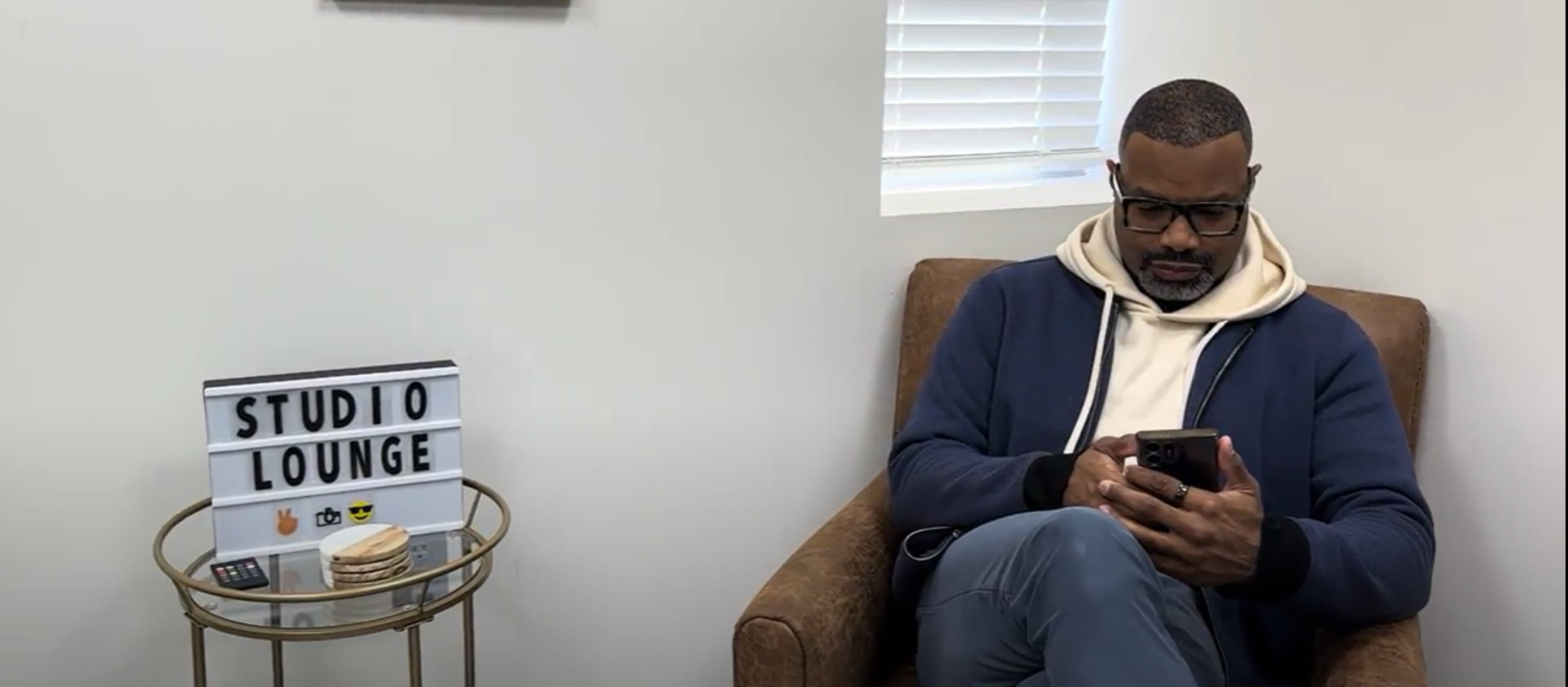 A man in a blue jacket and glasses sits in a chair while looking at his phone next to a sign that reads "STUDIO LOUNGE" on a small table.