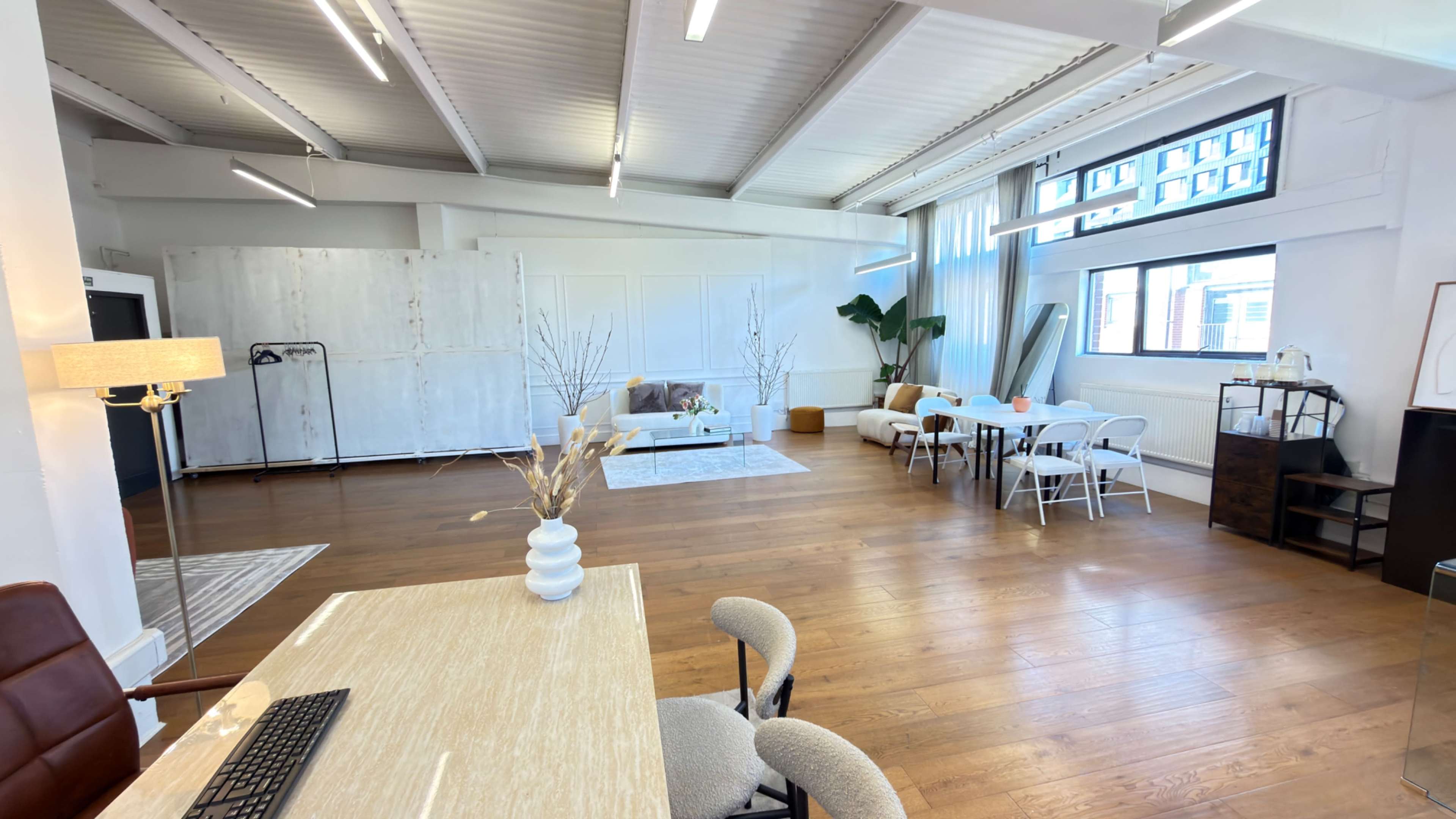 The image shows a spacious, well-lit interior featuring a mix of seating areas, a dining table, and decorative plants against a backdrop of large windows.