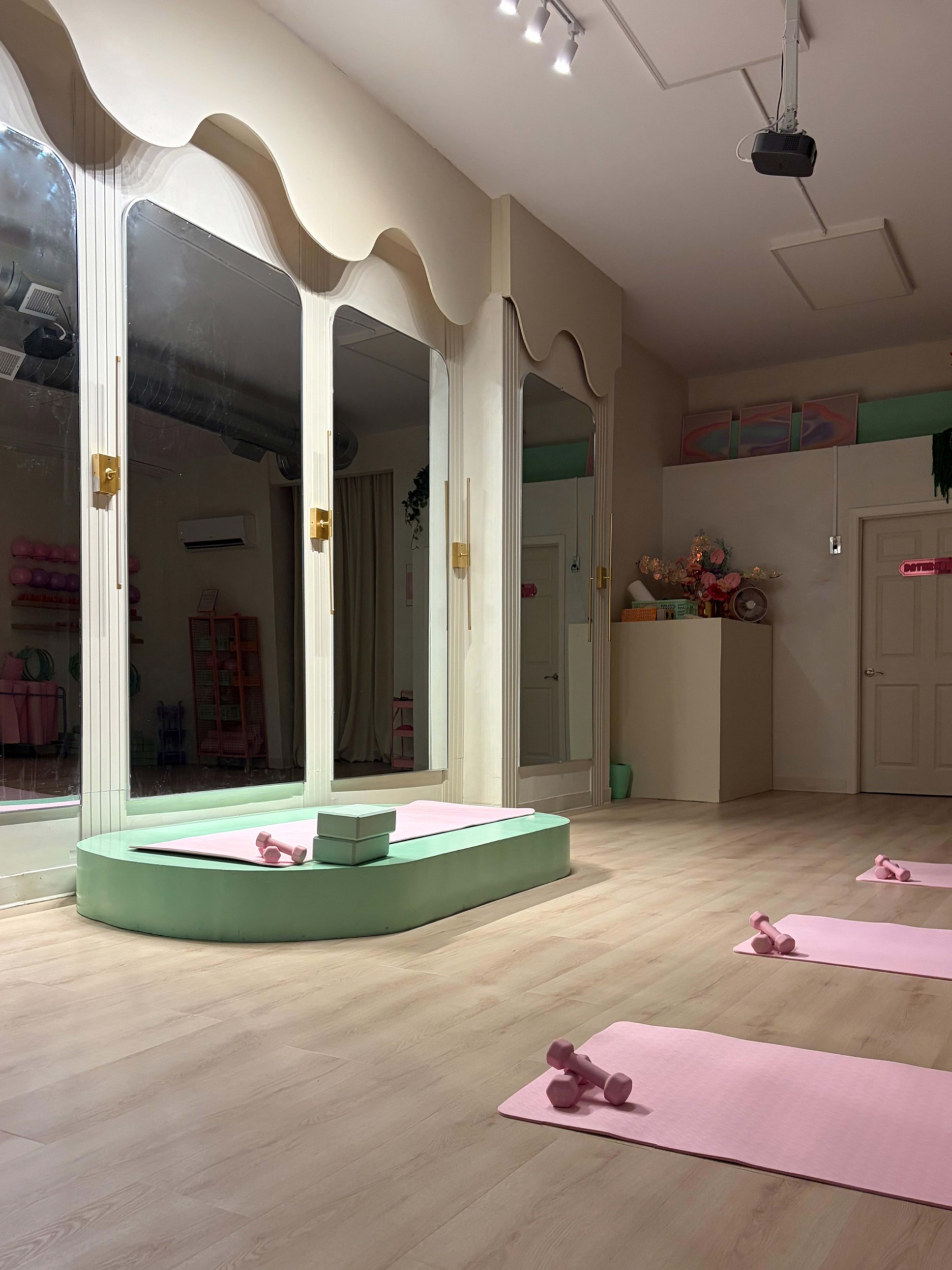 The image depicts a light-colored fitness studio featuring three large mirrors, a small platform with weights, and several exercise mats arranged on the floor.