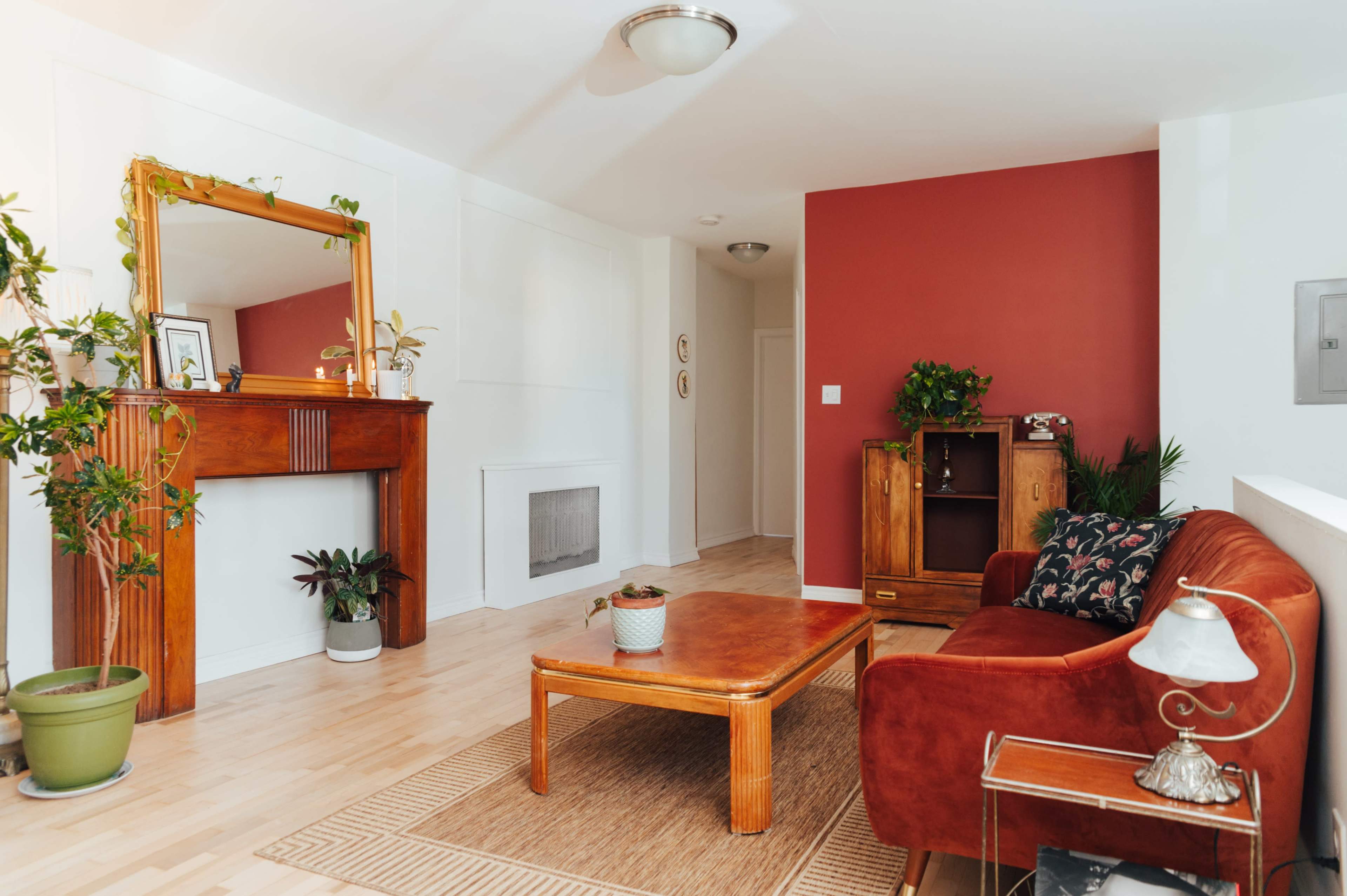 The room features a wooden fireplace mantel, a red accent wall, a brown couch, and a coffee table surrounded by plants.