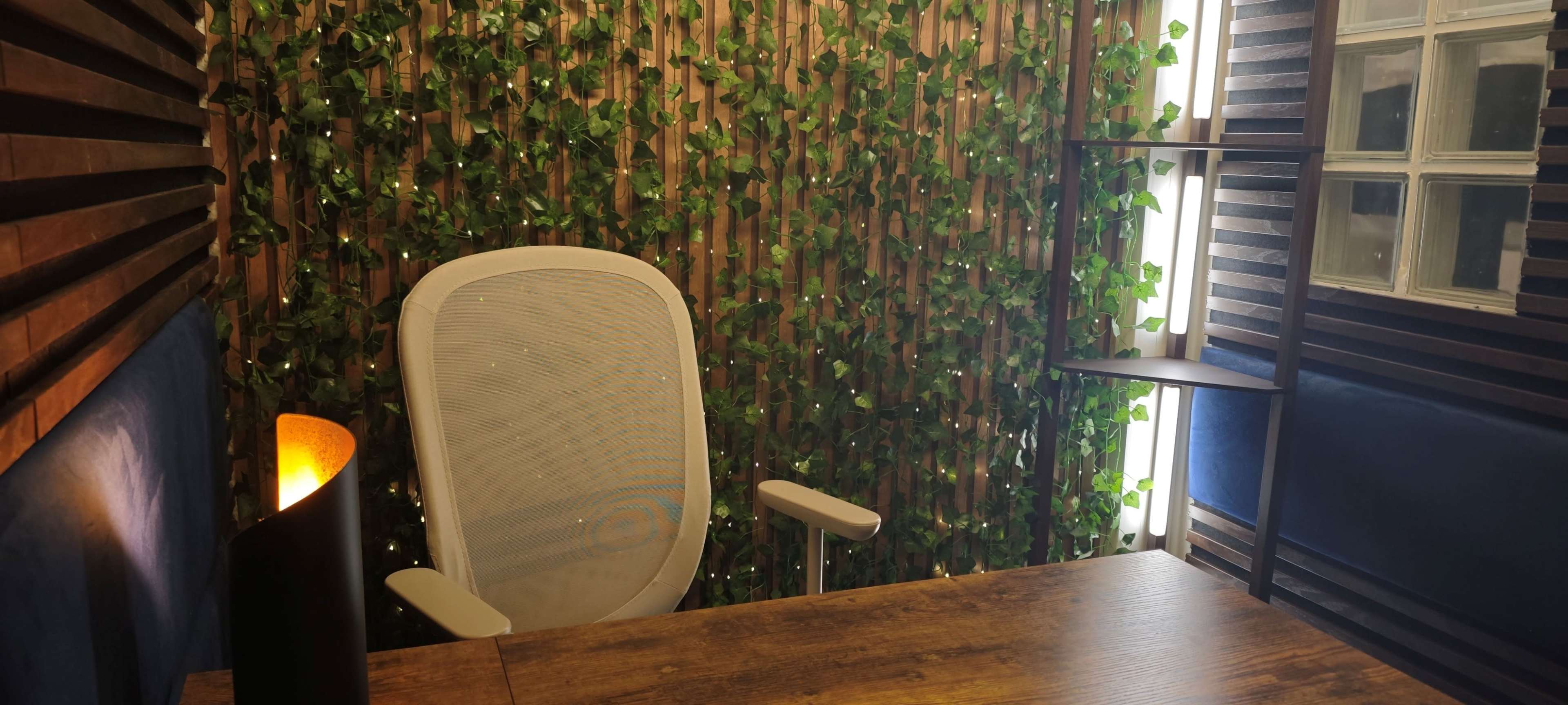 The image shows a modern office space featuring a wooden desk, a white ergonomic chair, and a backdrop of greenery along a vertical wooden panel.