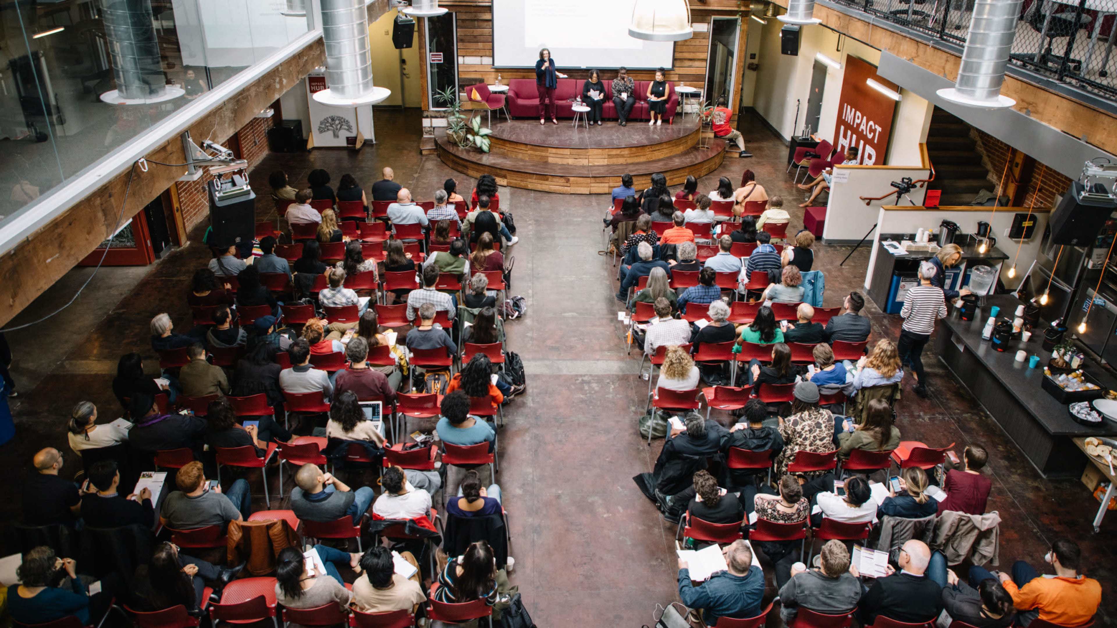 A large audience is seated in red chairs, facing a speaker at a presentation in a spacious venue with wooden accents.