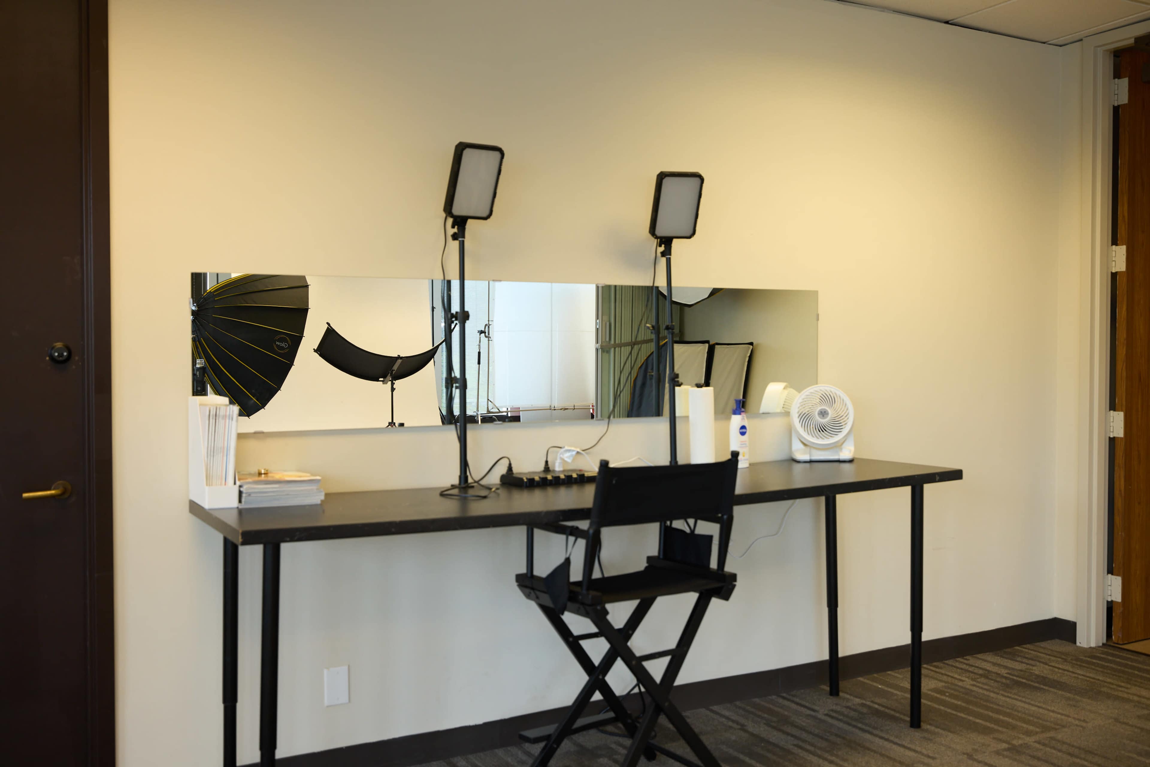 A simple makeup station is set up against a wall, featuring a long mirror, two lights, a fan, and a director's chair.