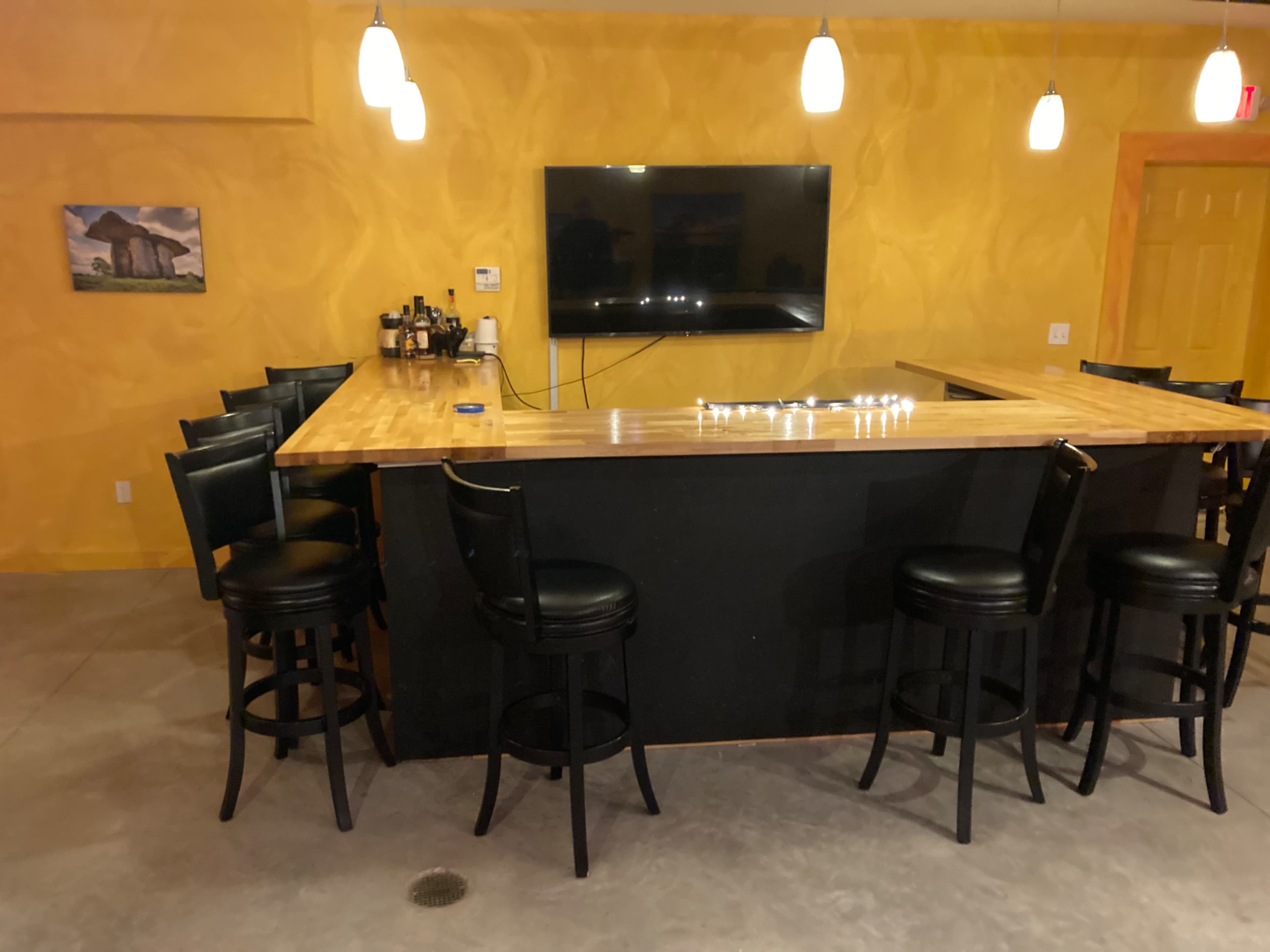 A bar setup with a wooden countertop, six black stools, a mounted television, and a yellow wall in a dimly lit room.