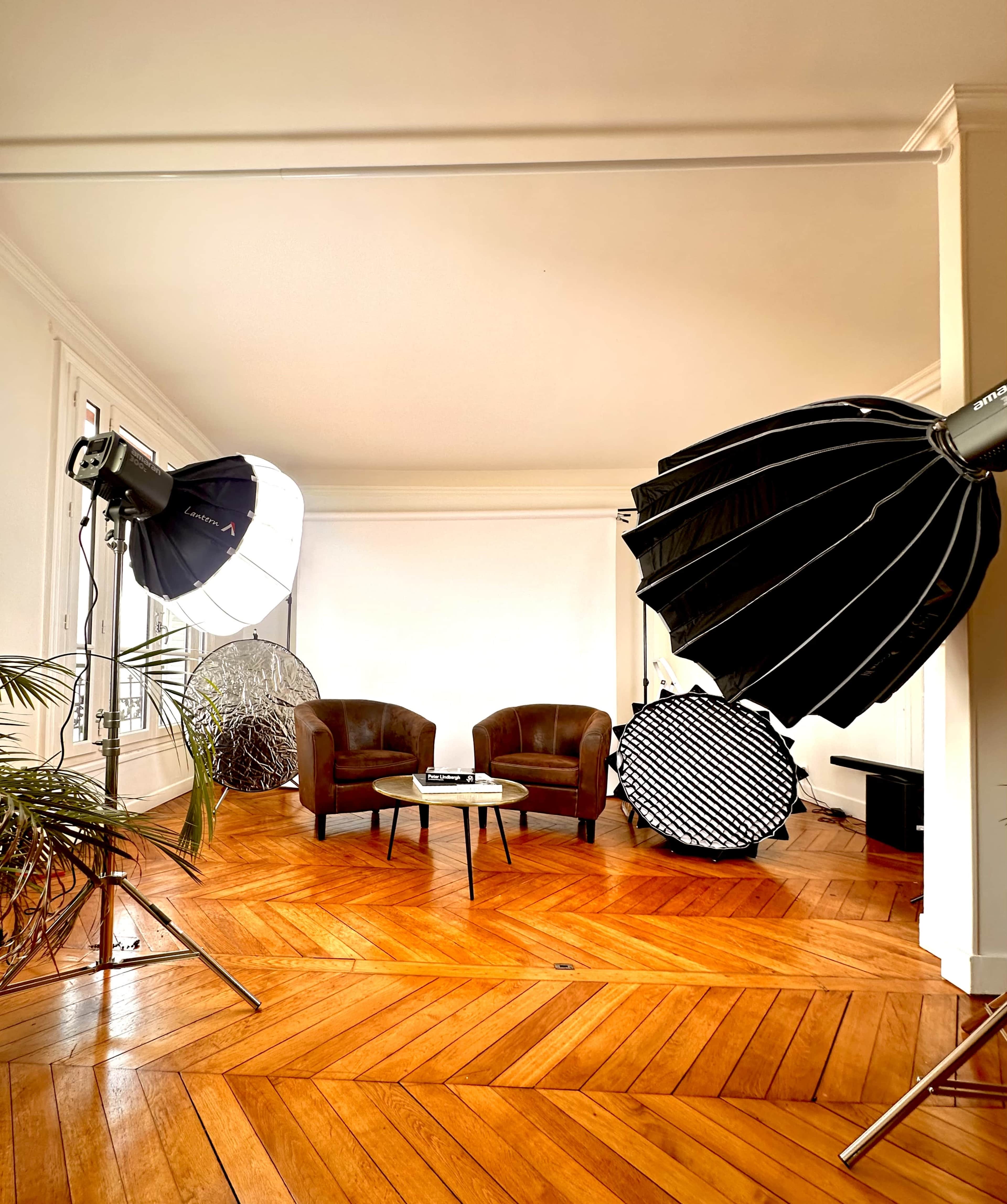 The image shows a photography studio with two brown chairs, a coffee table, and various lighting equipment set up on wooden flooring.
