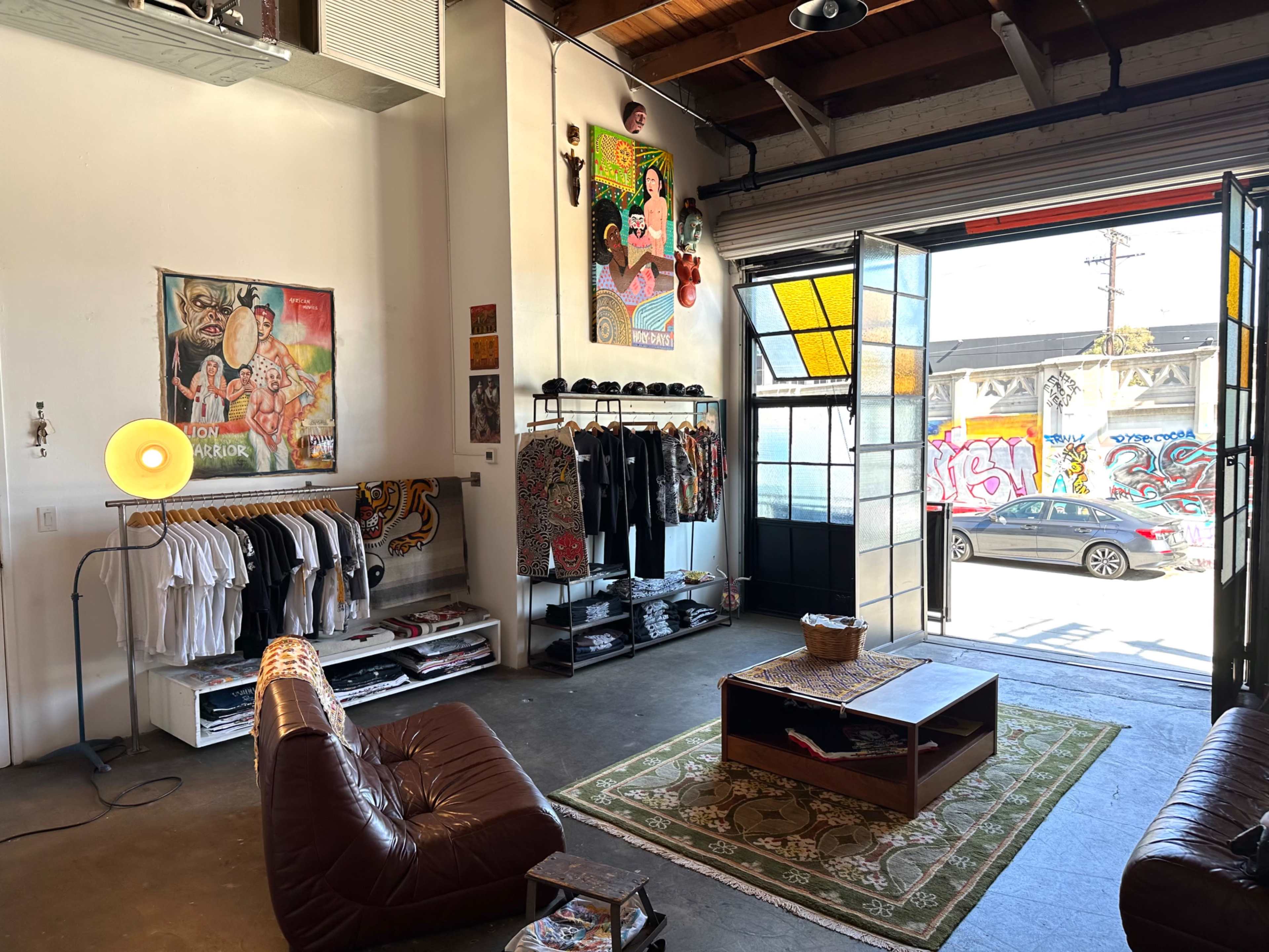 The image shows a contemporary clothing store interior with artwork on the walls, clothing racks filled with apparel, and an open door revealing a mural outside.