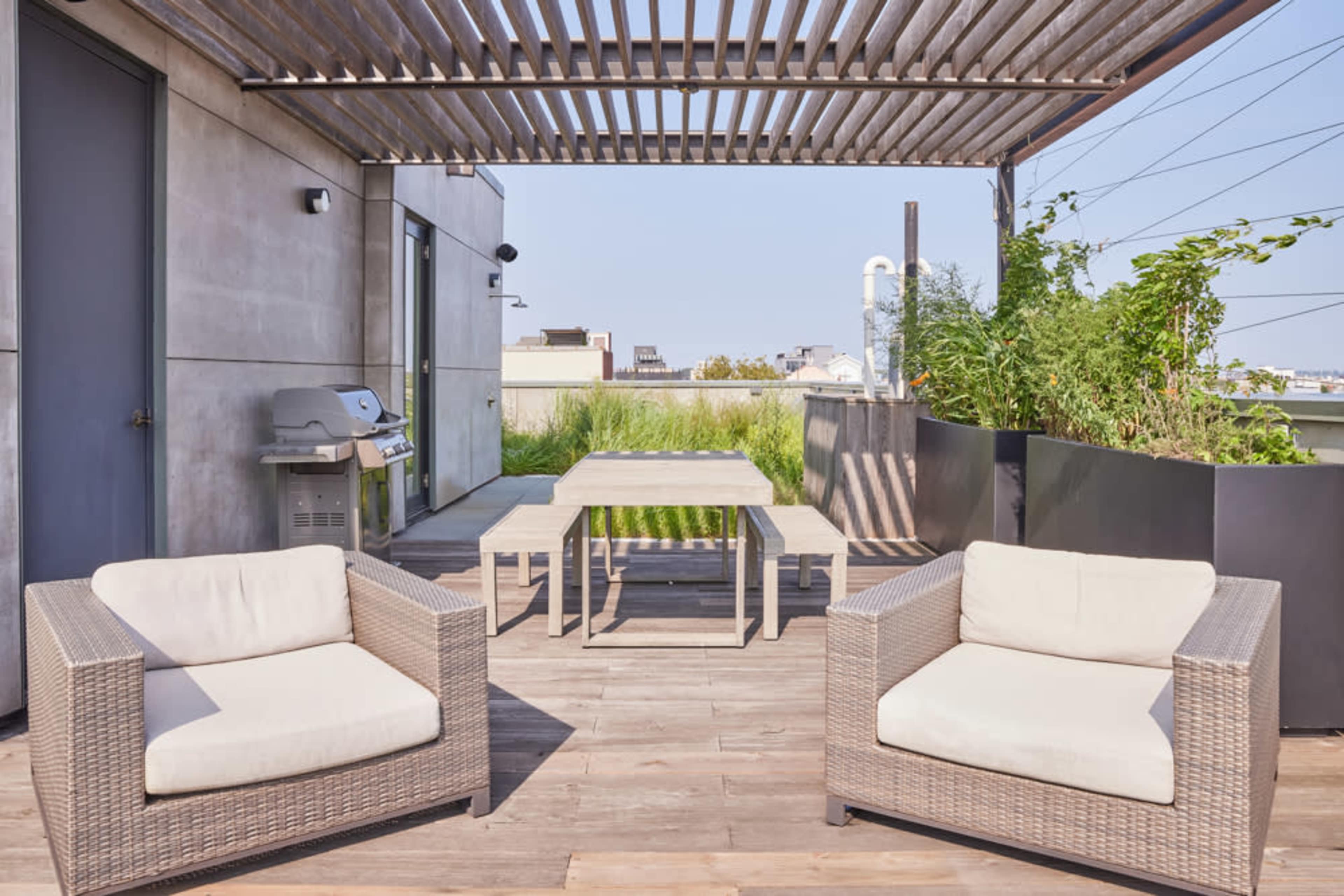 A modern rooftop terrace featuring two wicker armchairs, a wooden table, and a barbecue grill under a slatted pergola.