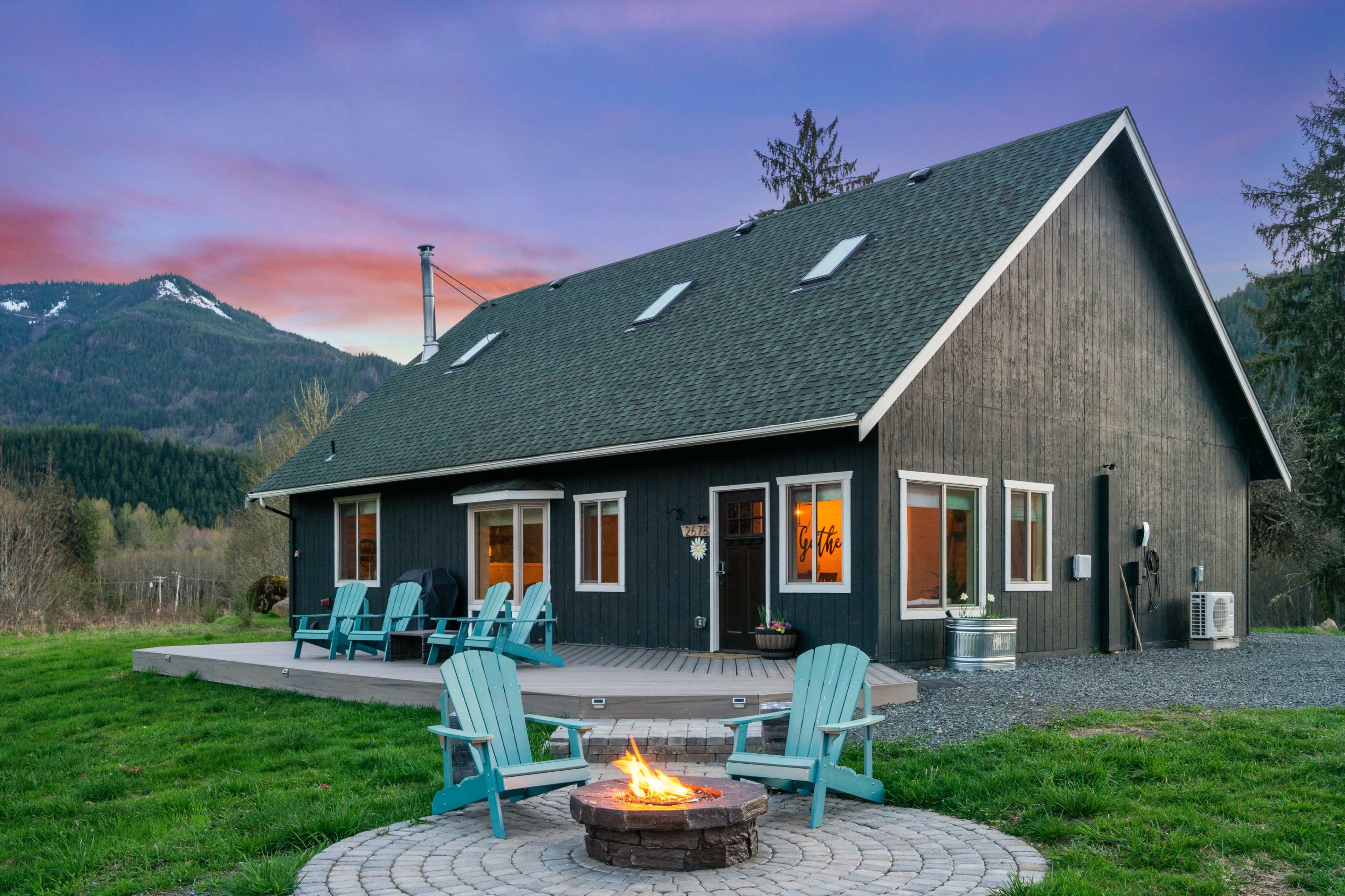 A modern black house with large windows and a deck features a stone fire pit and Adirondack chairs in a grassy area near mountains.