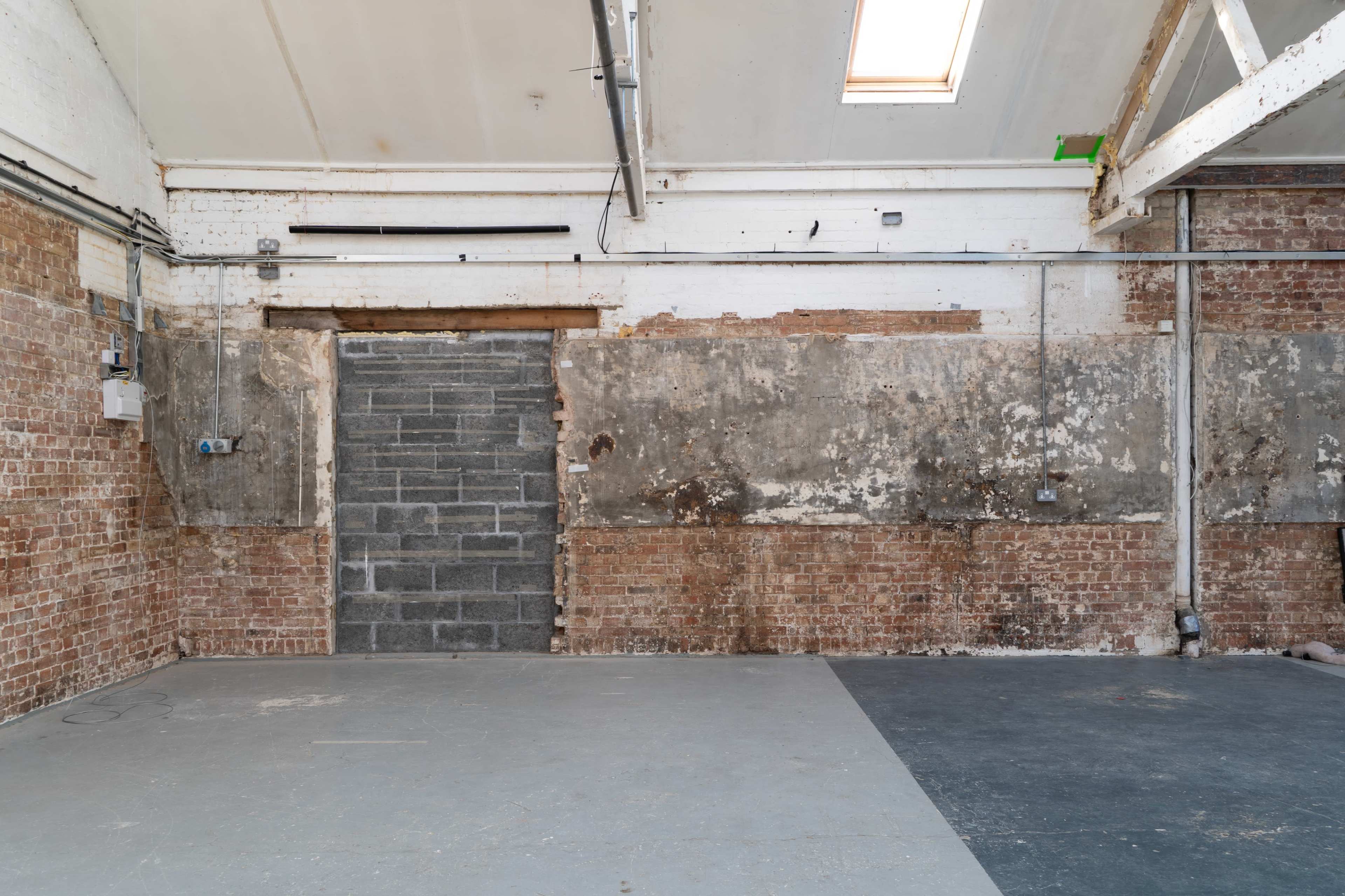 The image shows an industrial interior with a partially bricked wall, concrete floor, and exposed beams.