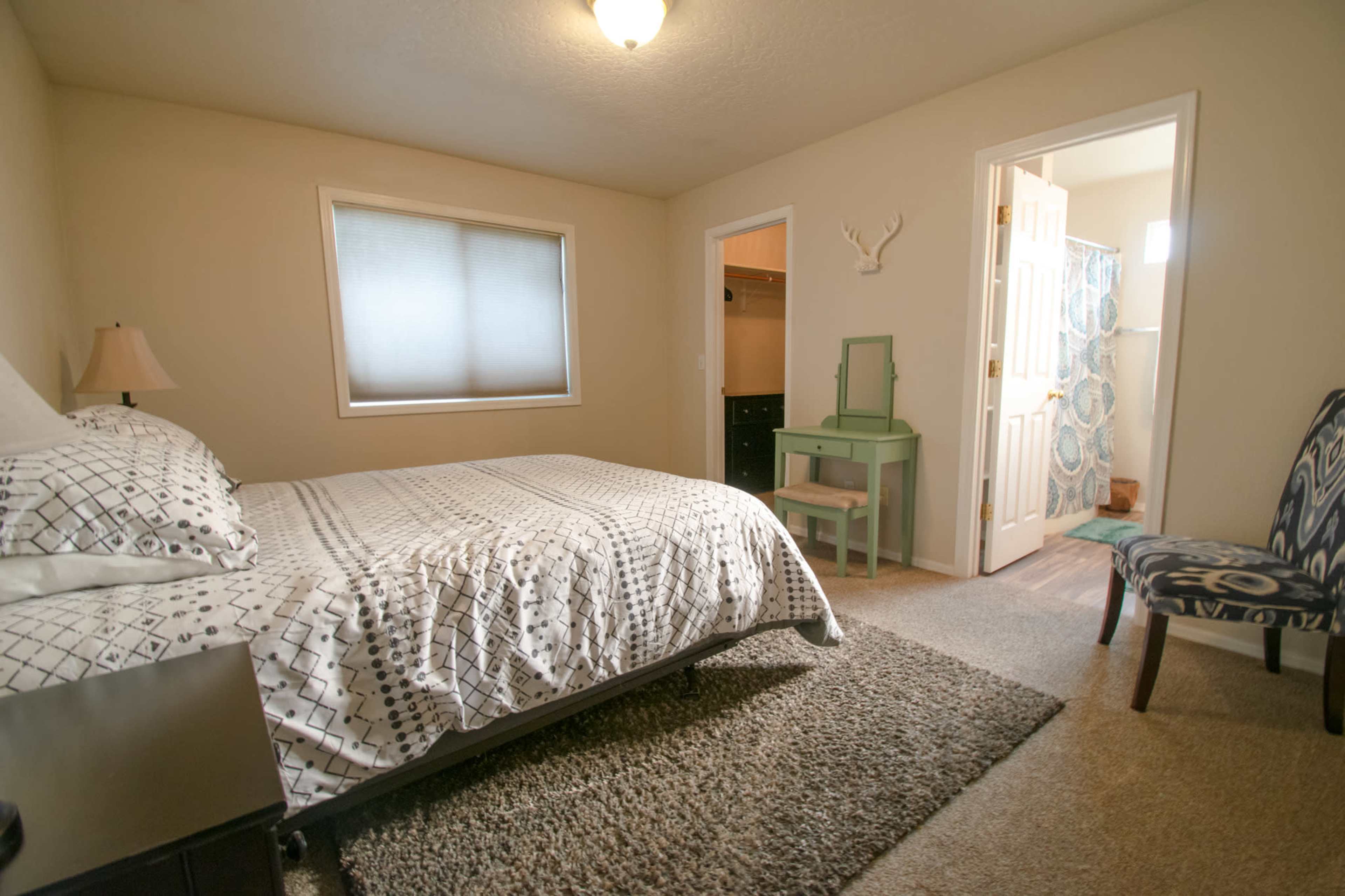 A tidy bedroom featuring a bed with a patterned comforter, a small green table, and a chair, with a closet visible in the background.