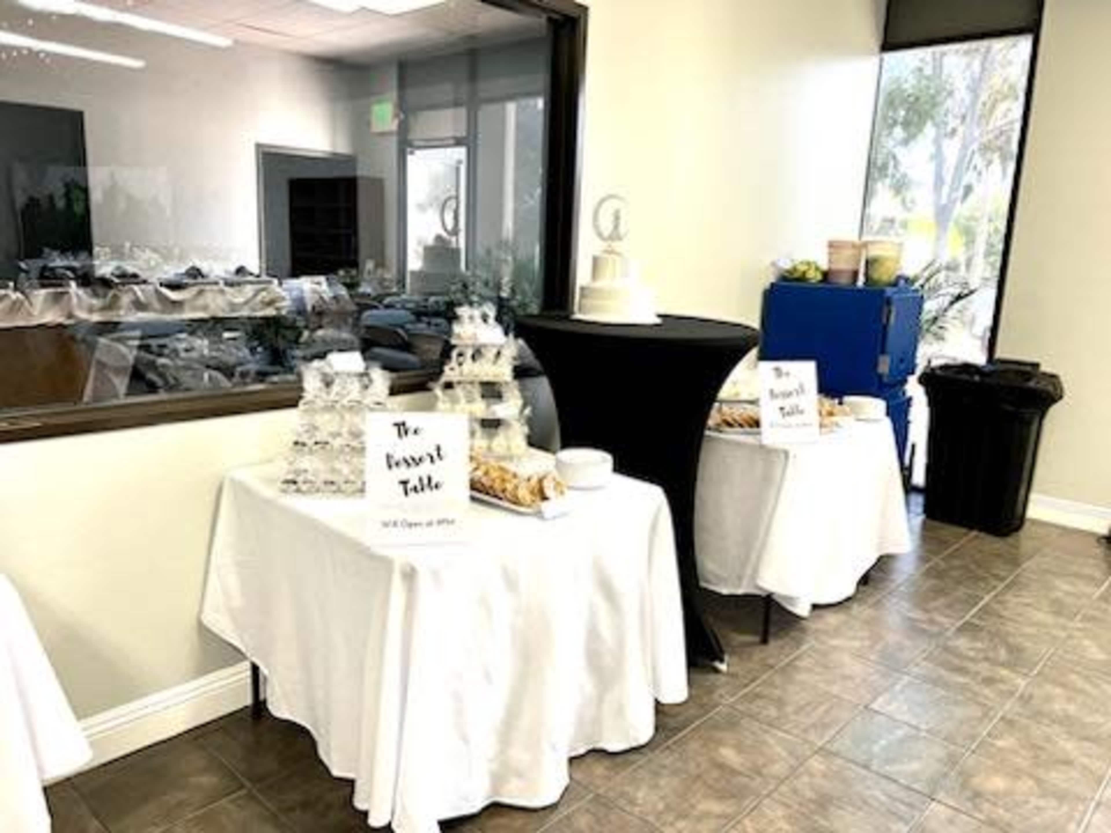 A room with two tables set up for a buffet, featuring clear dessert cups, pastries, and a wedding cake, along with signage labeling "The Dessert Table."