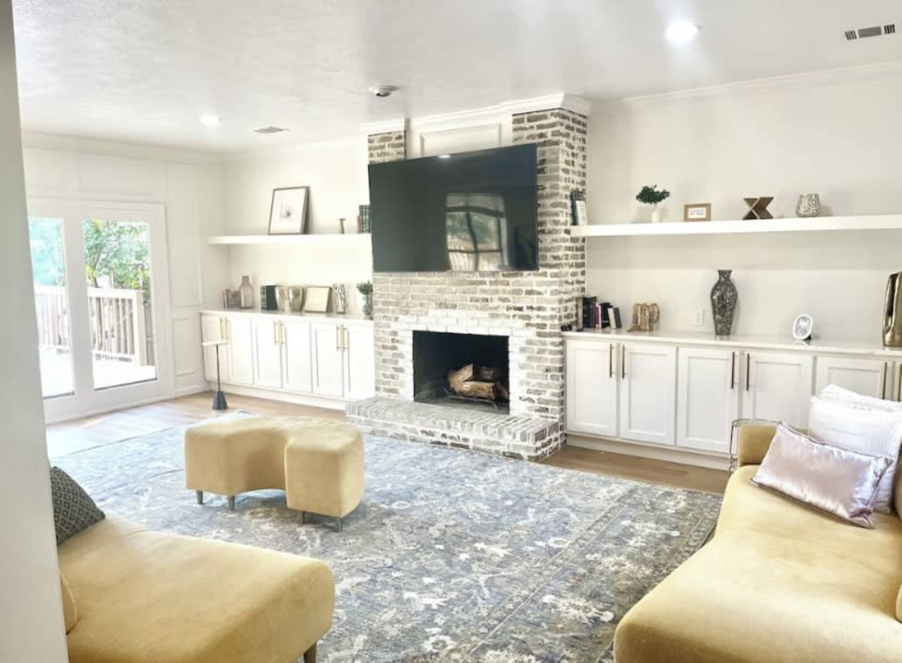 The living room features a brick fireplace, a large television mounted on the wall, and shelves filled with decorative items, alongside two couches and a patterned blue rug.