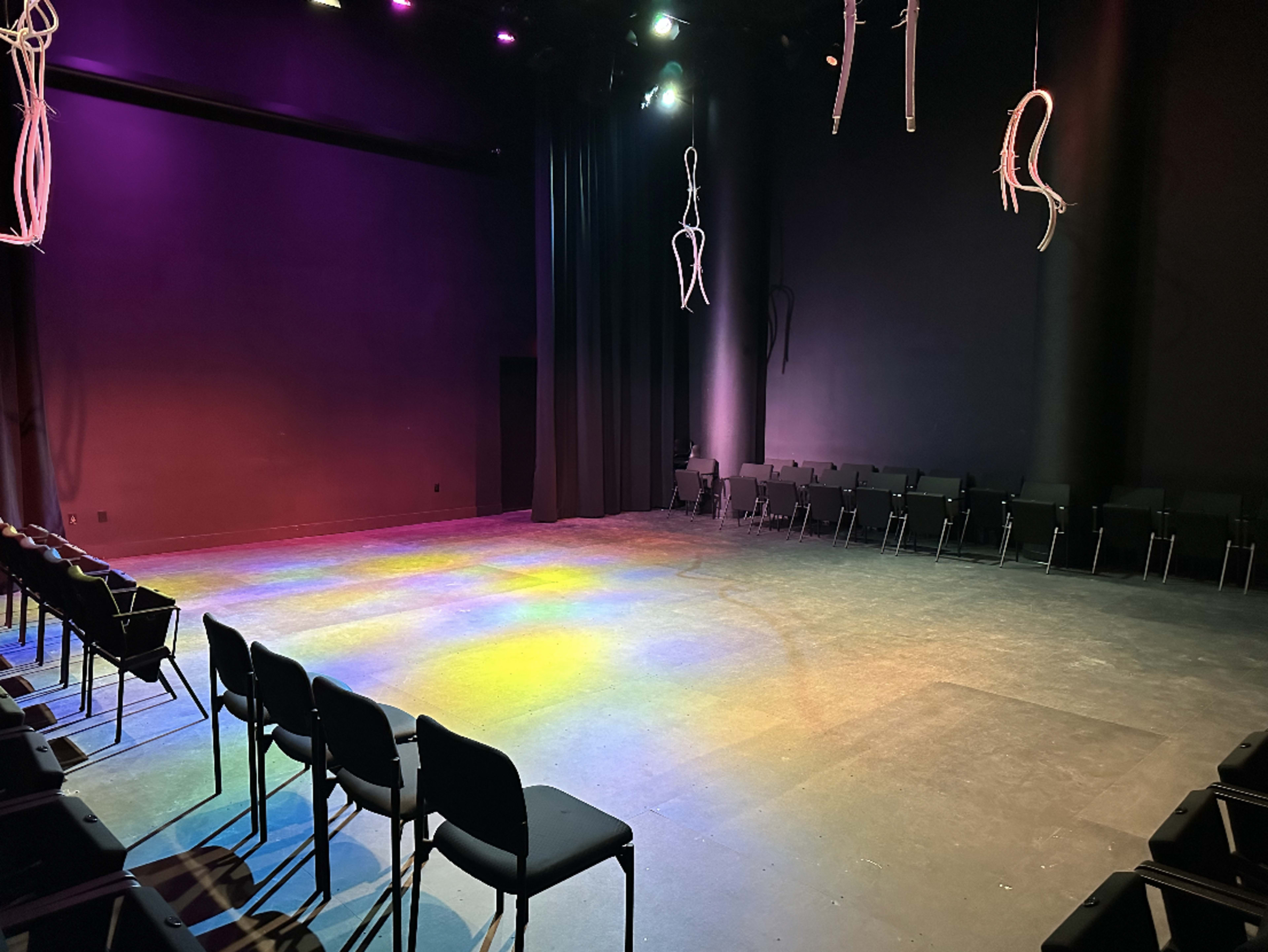 A dimly lit stage space features rows of black chairs arranged in a semicircle, with colorful lights casting patterns on the floor and suspended ropes hanging from the ceiling.