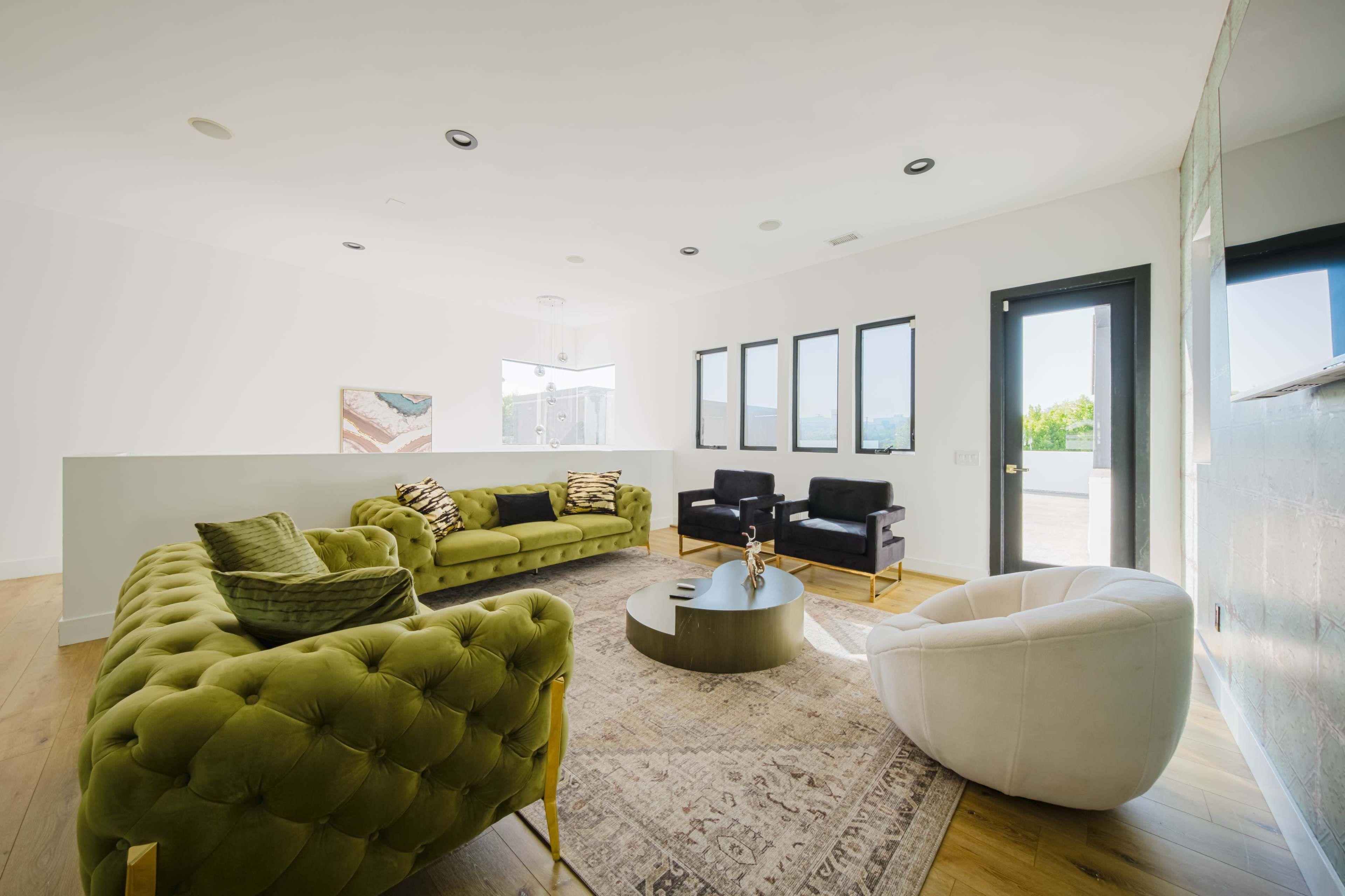 The image shows a modern living room featuring two green tufted sofas, two black armchairs, and a white circular chair arranged around a round coffee table on a patterned rug.