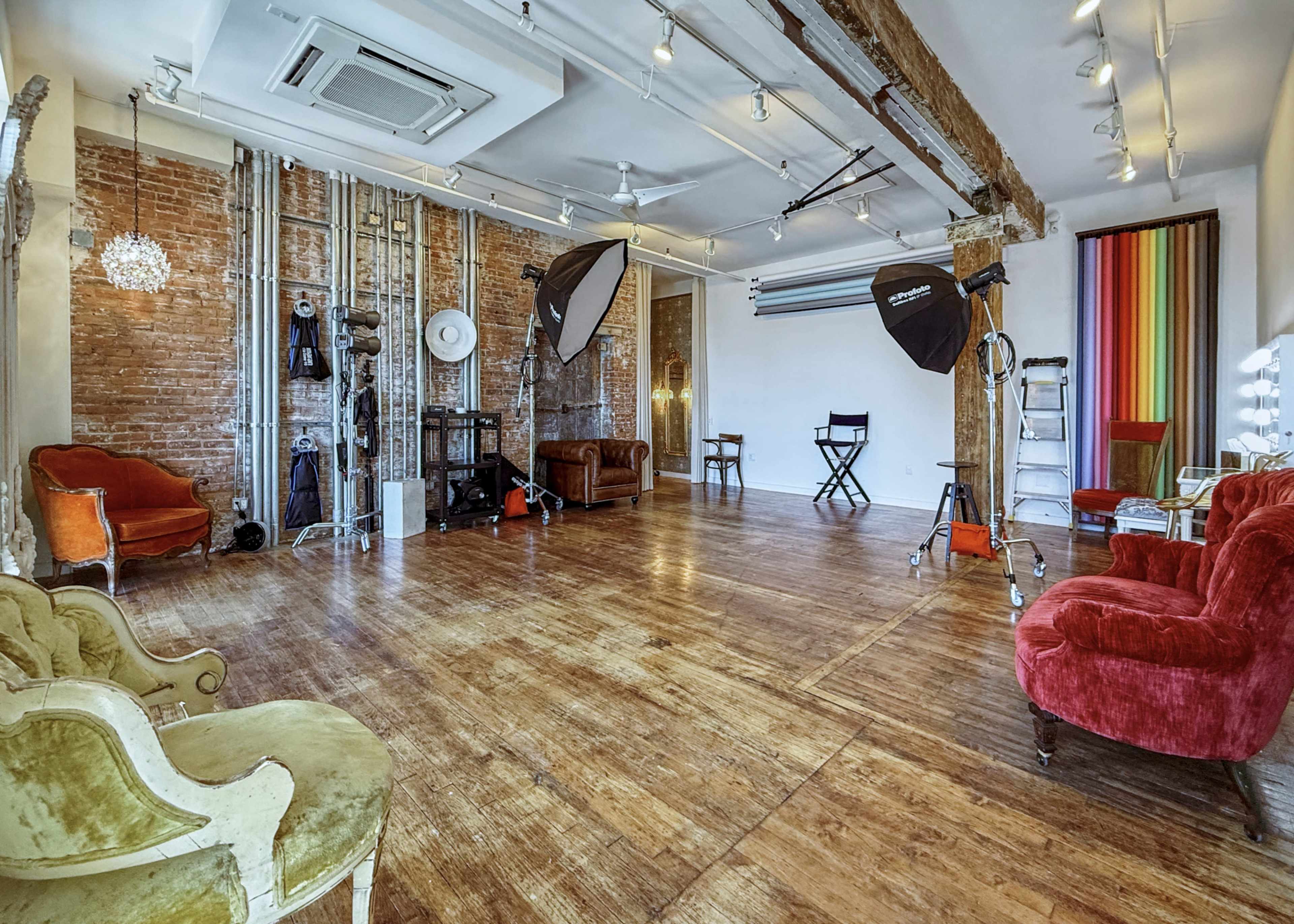 The image shows a spacious studio with wooden floors, exposed brick walls, and various photography equipment, including softbox lights and a makeup vanity.