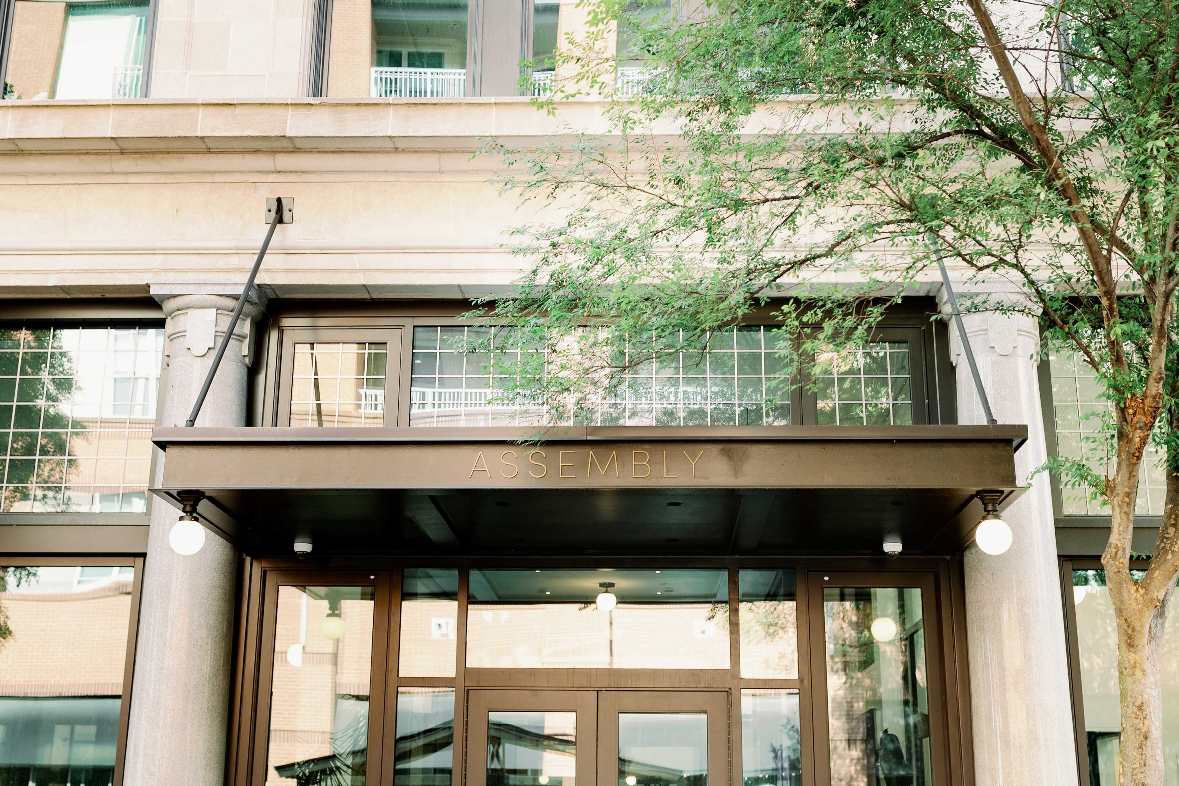 The image shows the entrance of a building with the word "ASSEMBLY" prominently displayed above the door, framed by large windows and supported by columns.