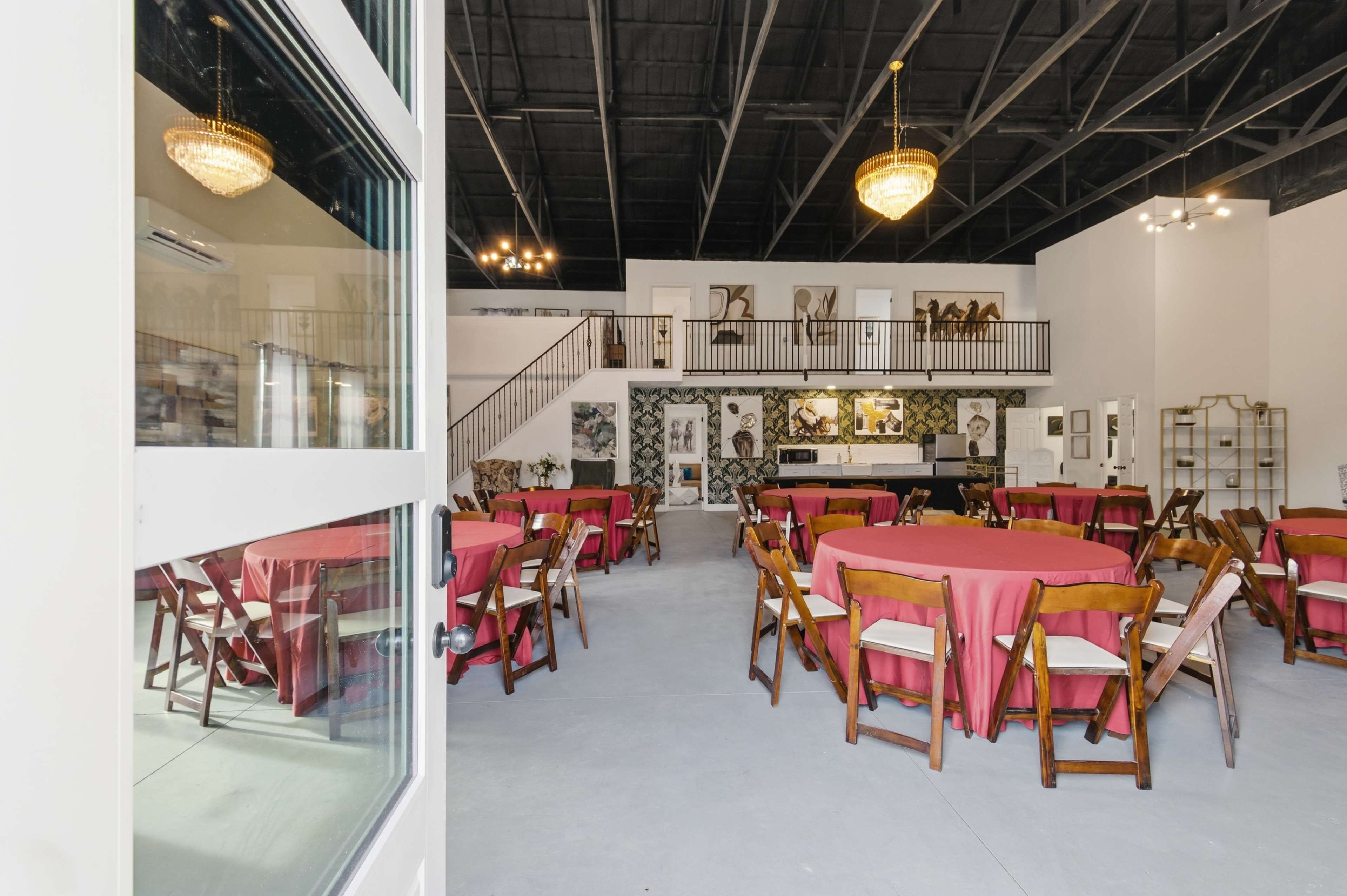 The image shows a spacious event venue with round tables dressed in red tablecloths and decorative wall art, illuminated by chandeliers.