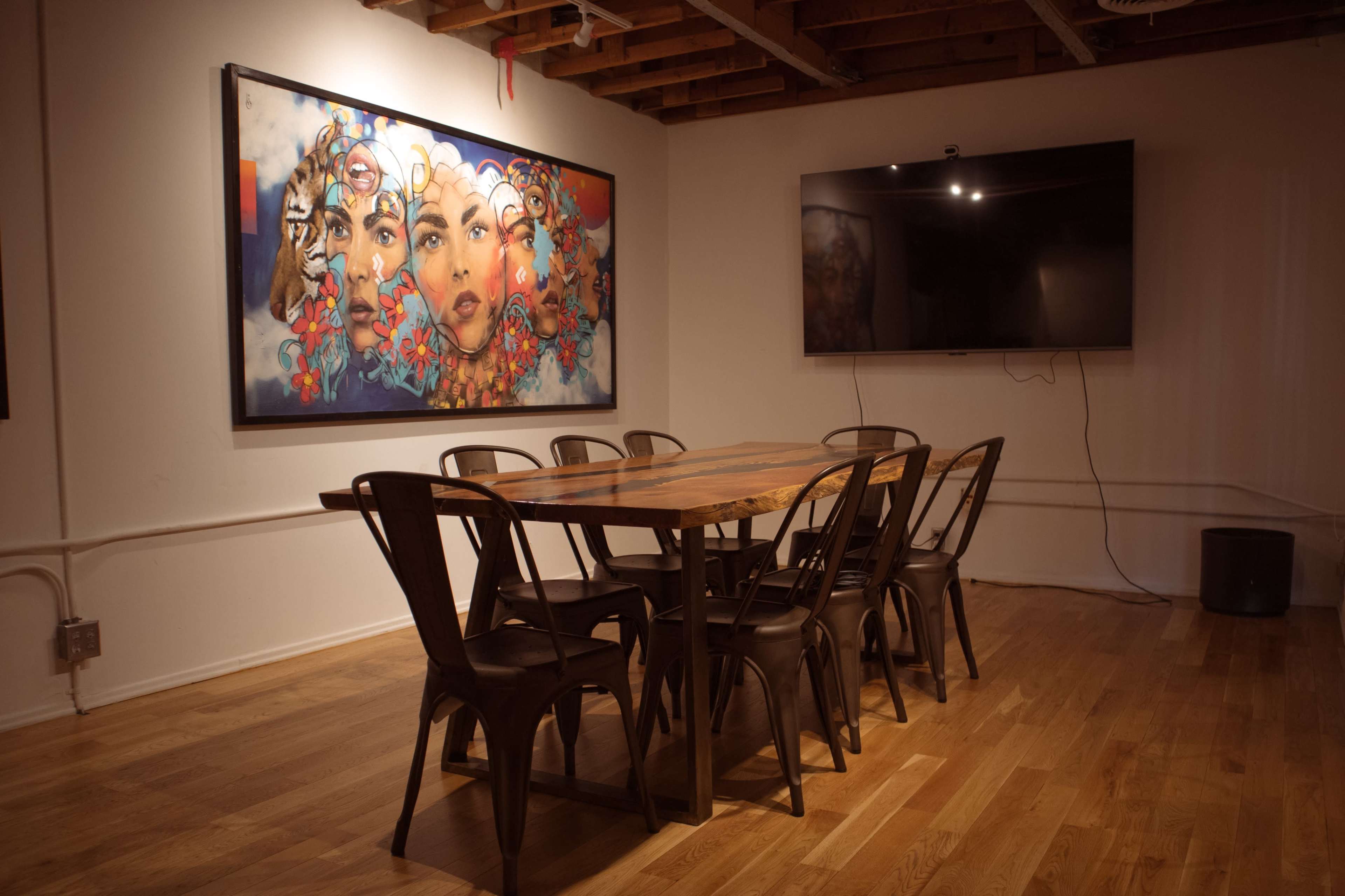A wooden dining table with eight metal chairs is set in a room featuring a large colorful mural on one wall and a flat-screen TV on another.
