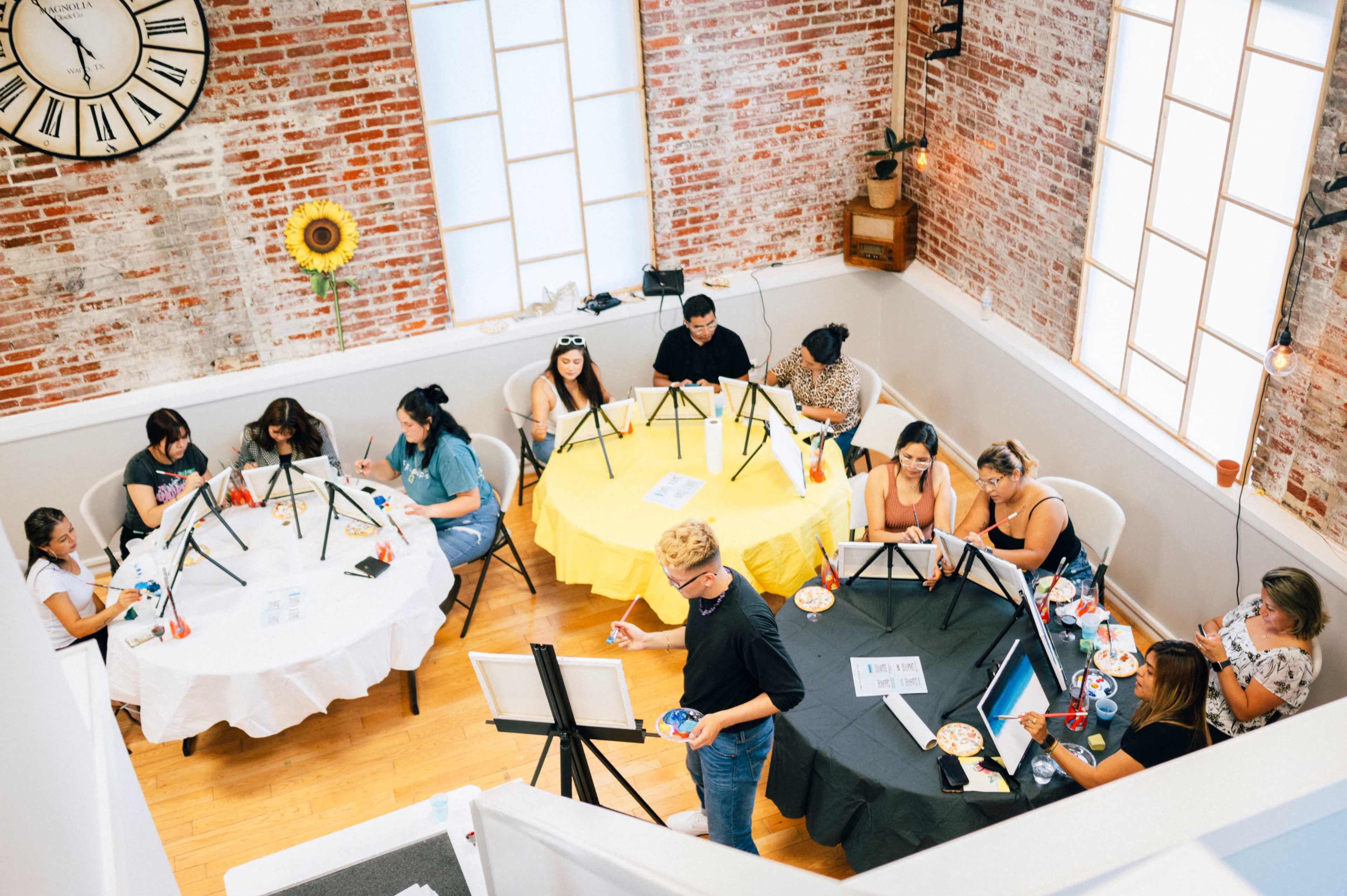 A group of people participates in an art painting class, seated at tables with easels and paint supplies in a bright, brick-walled room.