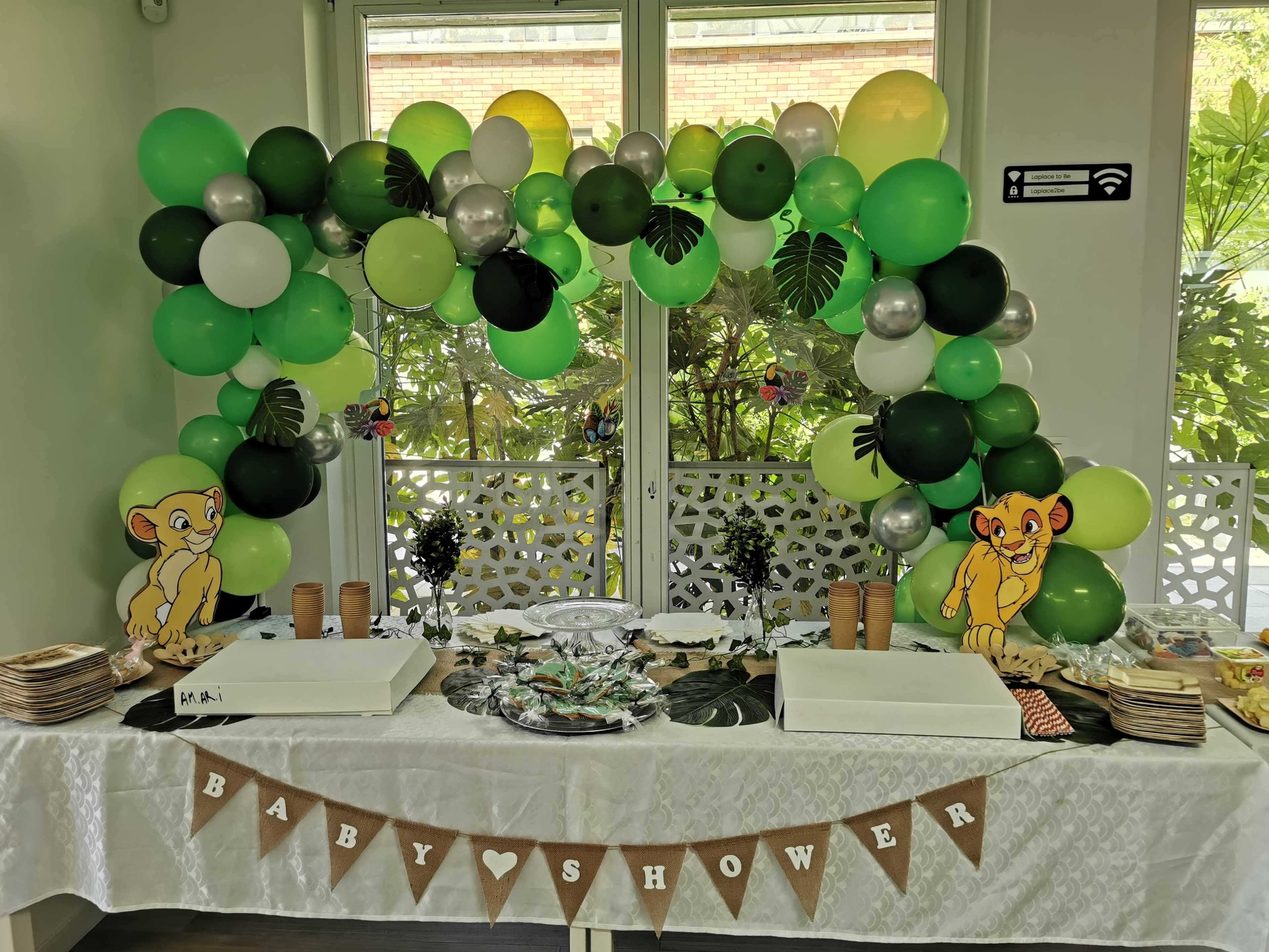 A baby shower table is decorated with green and black balloons, featuring a jungle theme with cartoon lion characters and a "BABY SHOWER" banner.