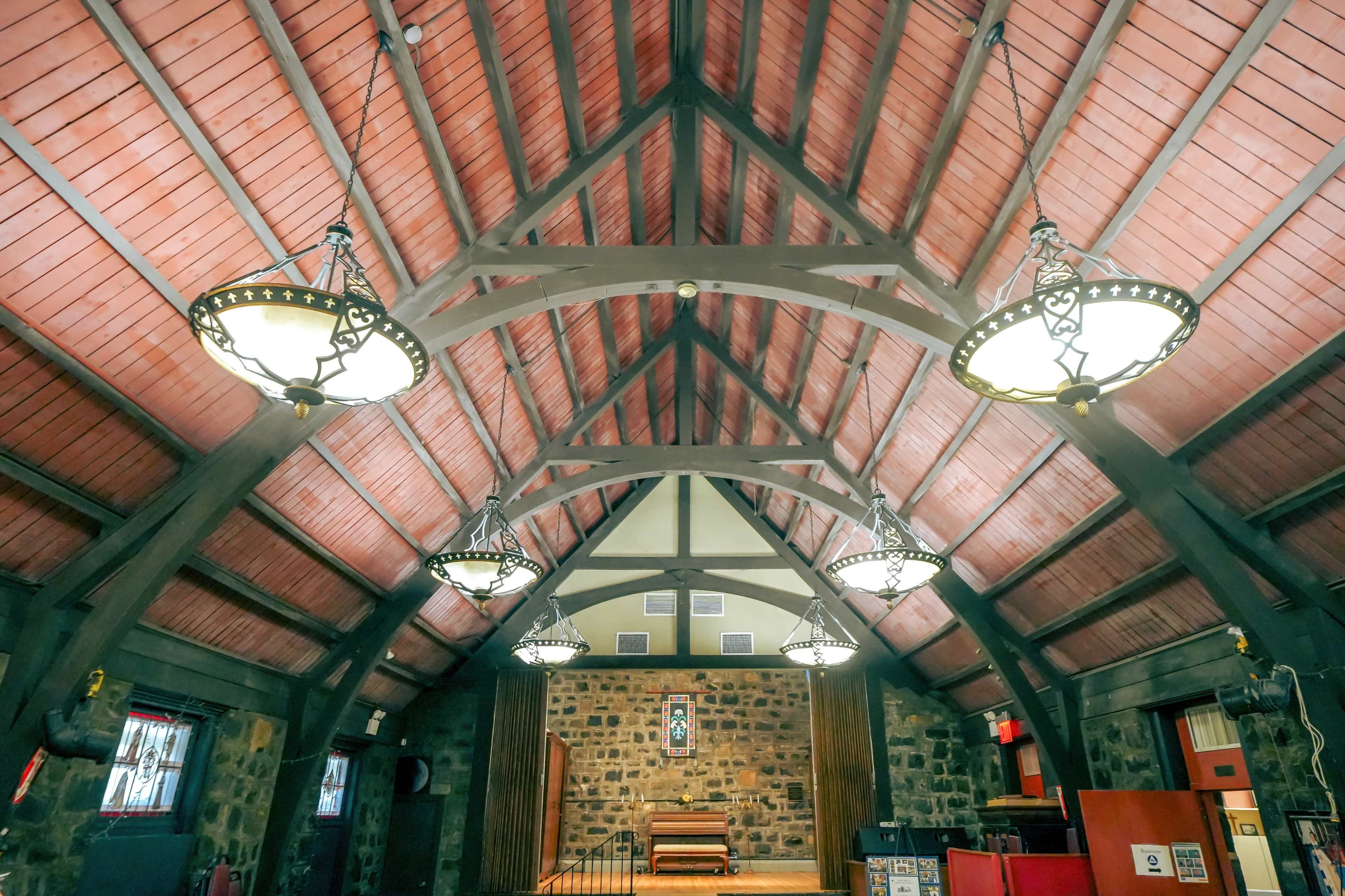 The interior of a building with a high wooden ceiling and multiple hanging light fixtures, featuring stone walls and a wooden stage at one end.