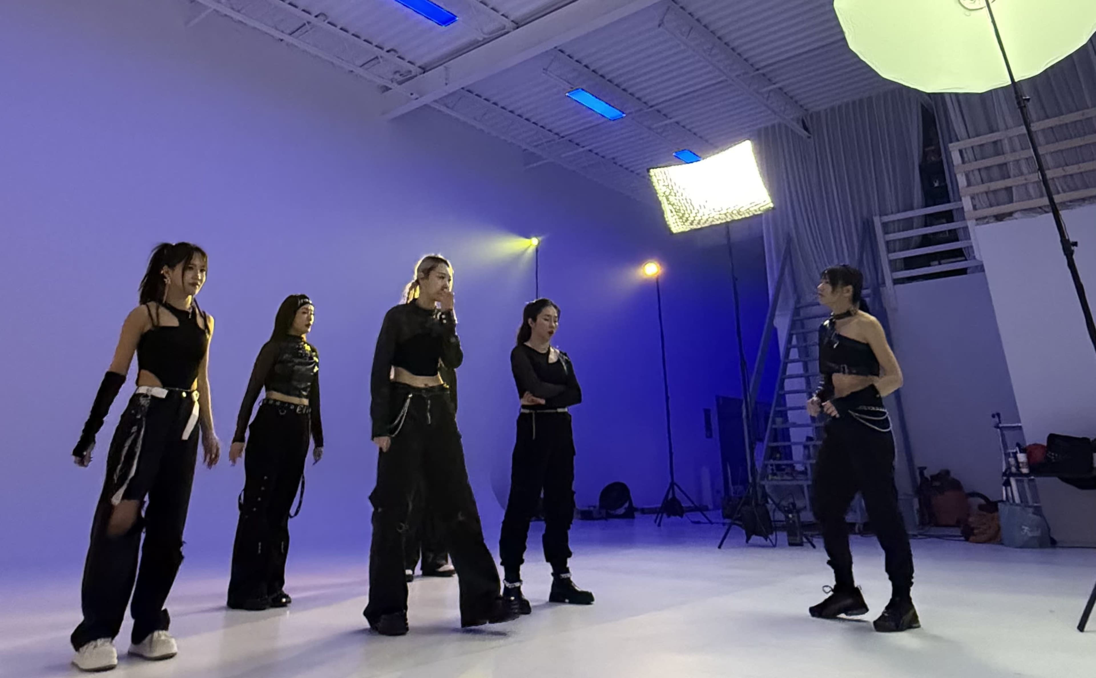 A group of dancers in black outfits rehearses under studio lights in a spacious dance studio.