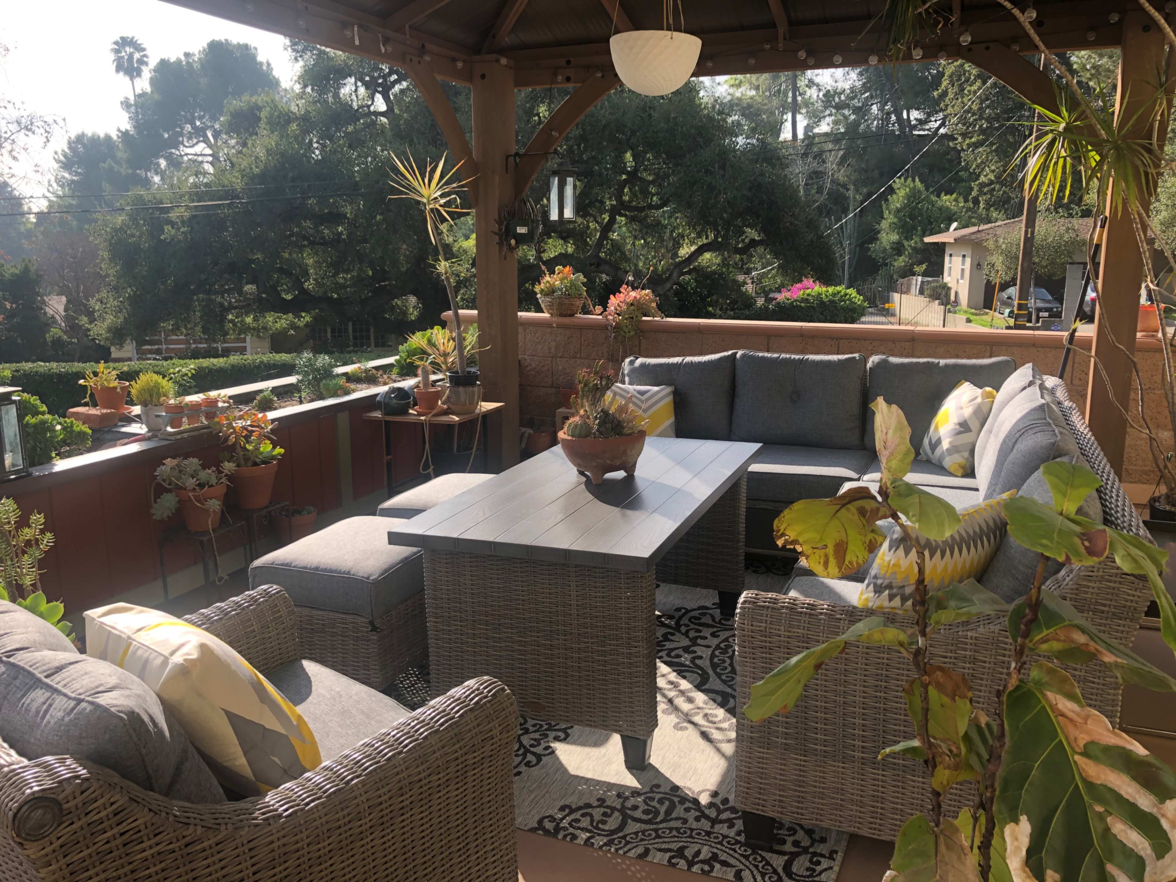 A shaded patio area features a sectional sofa and a coffee table surrounded by potted plants and decorative furnishings.