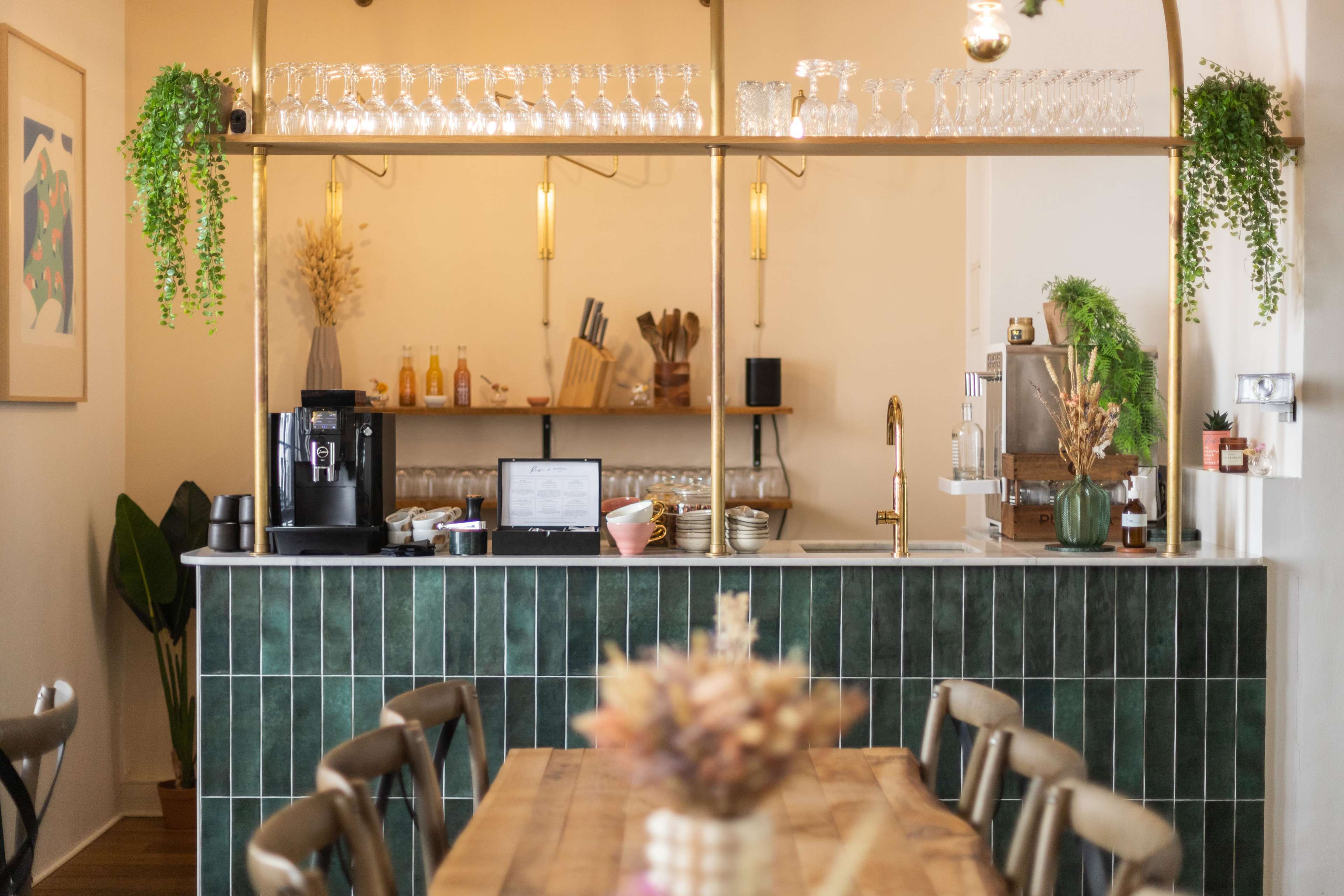 A coffee shop interior features a bar with glassware neatly arranged above, a coffee machine, and a green tiled counter.