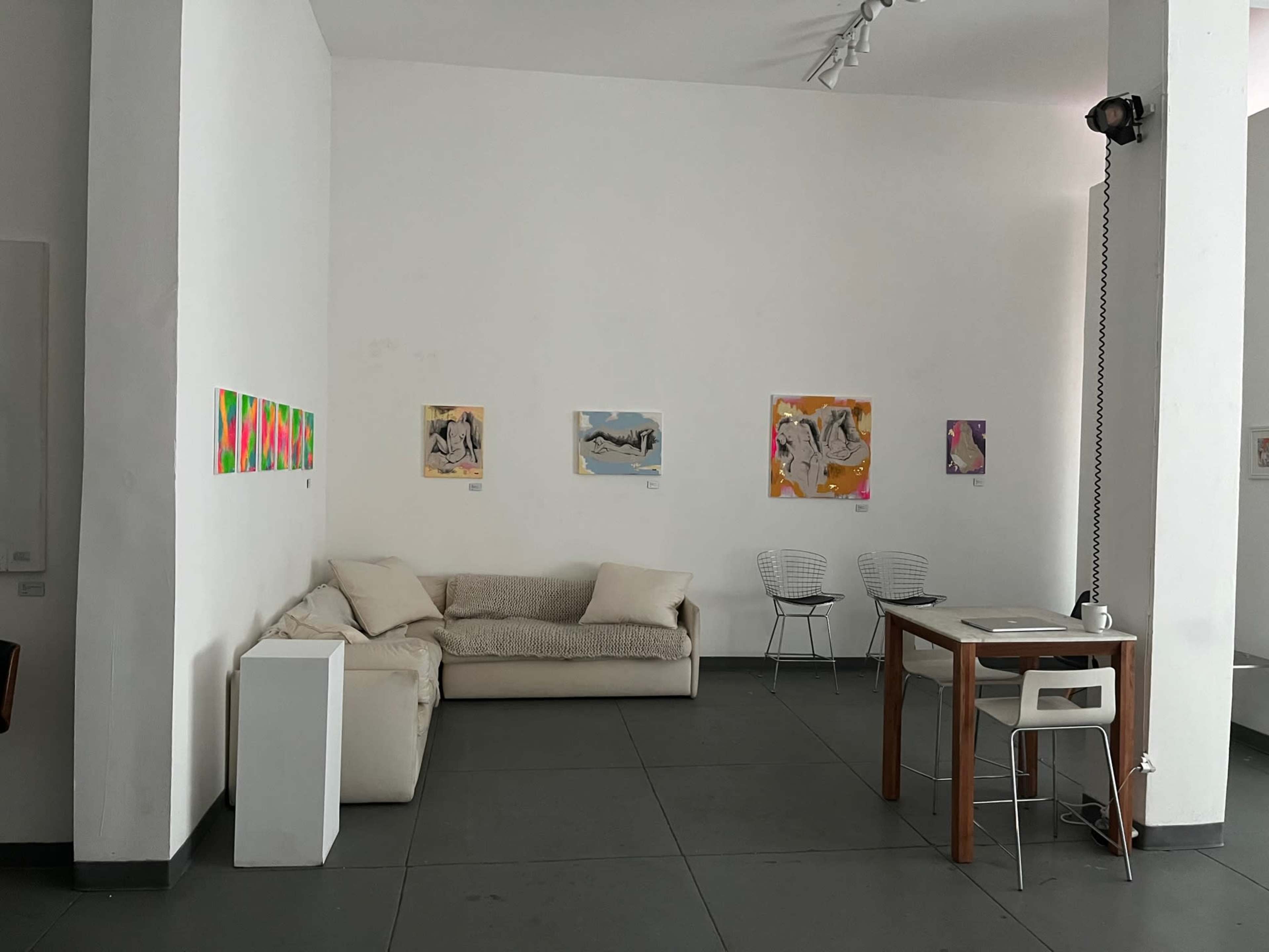 A minimalist gallery space with white walls, a light-colored sofa, a wooden table, and several colorful artworks hanging on the walls.