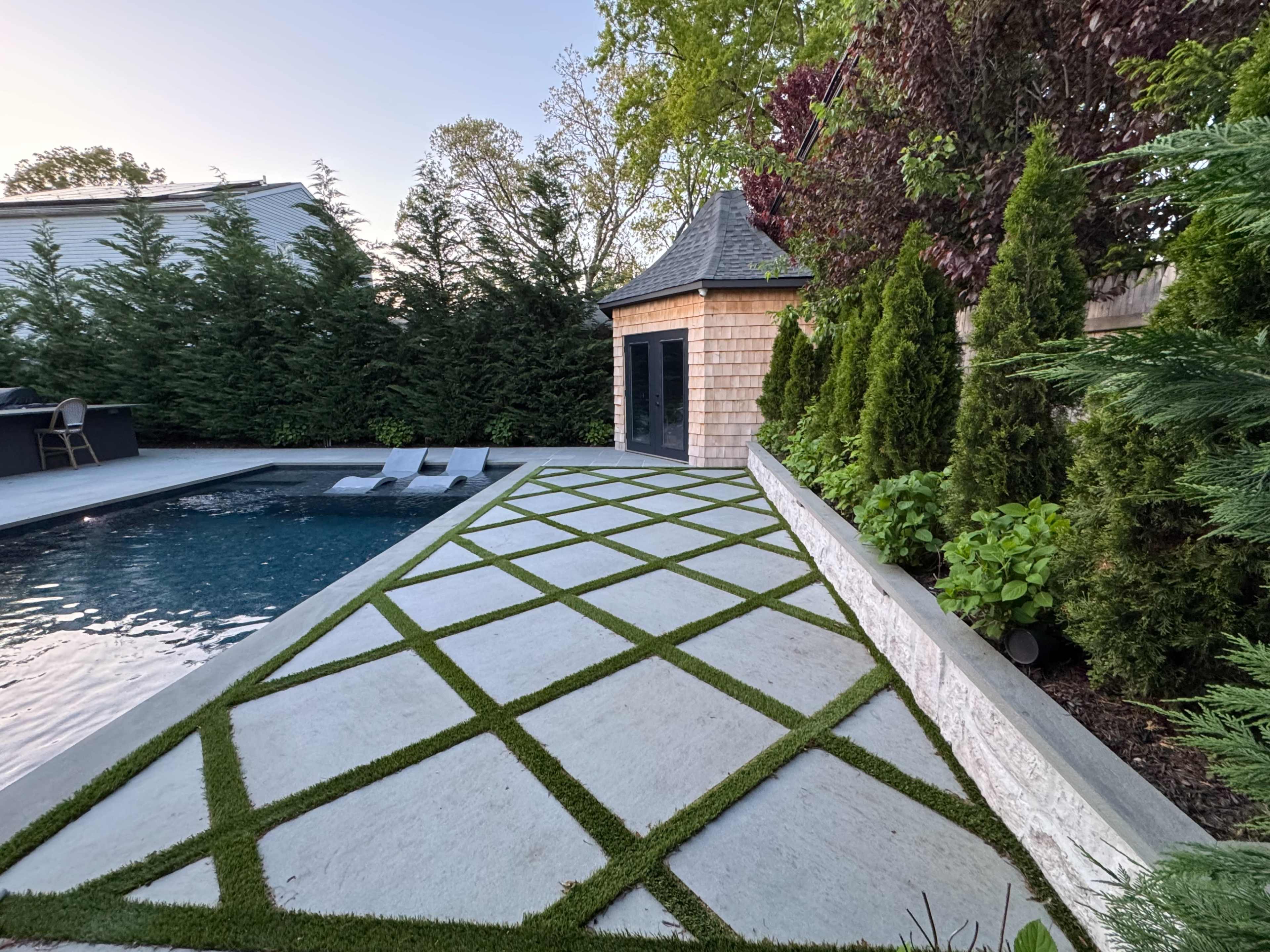 The image shows a sleek outdoor pool area with a stone pathway featuring a geometric pattern of grass inlays, bordered by lush greenery and a nearby building.