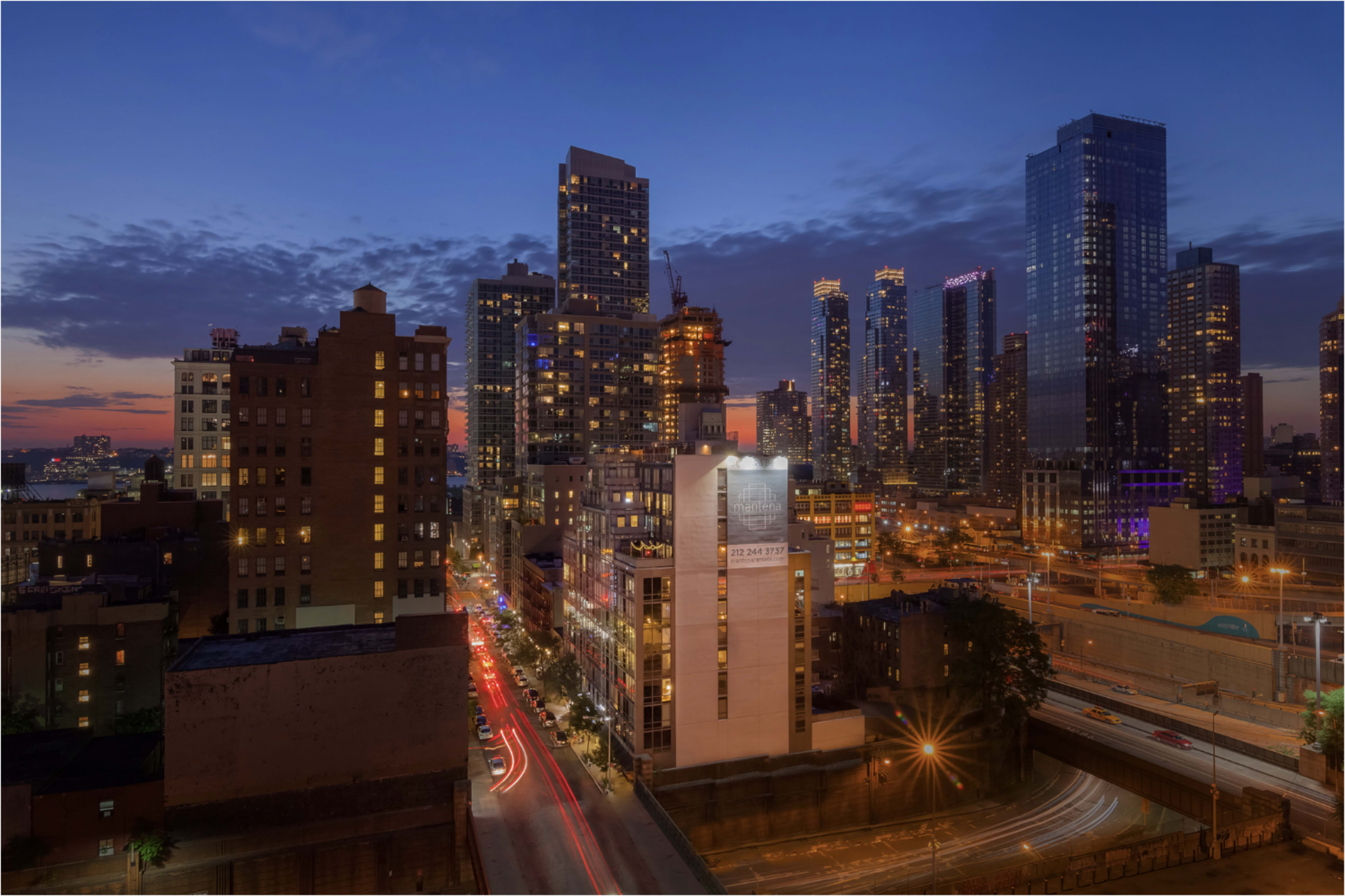 The image shows a city skyline at dusk, with illuminated buildings and streets, and a highway running beneath.