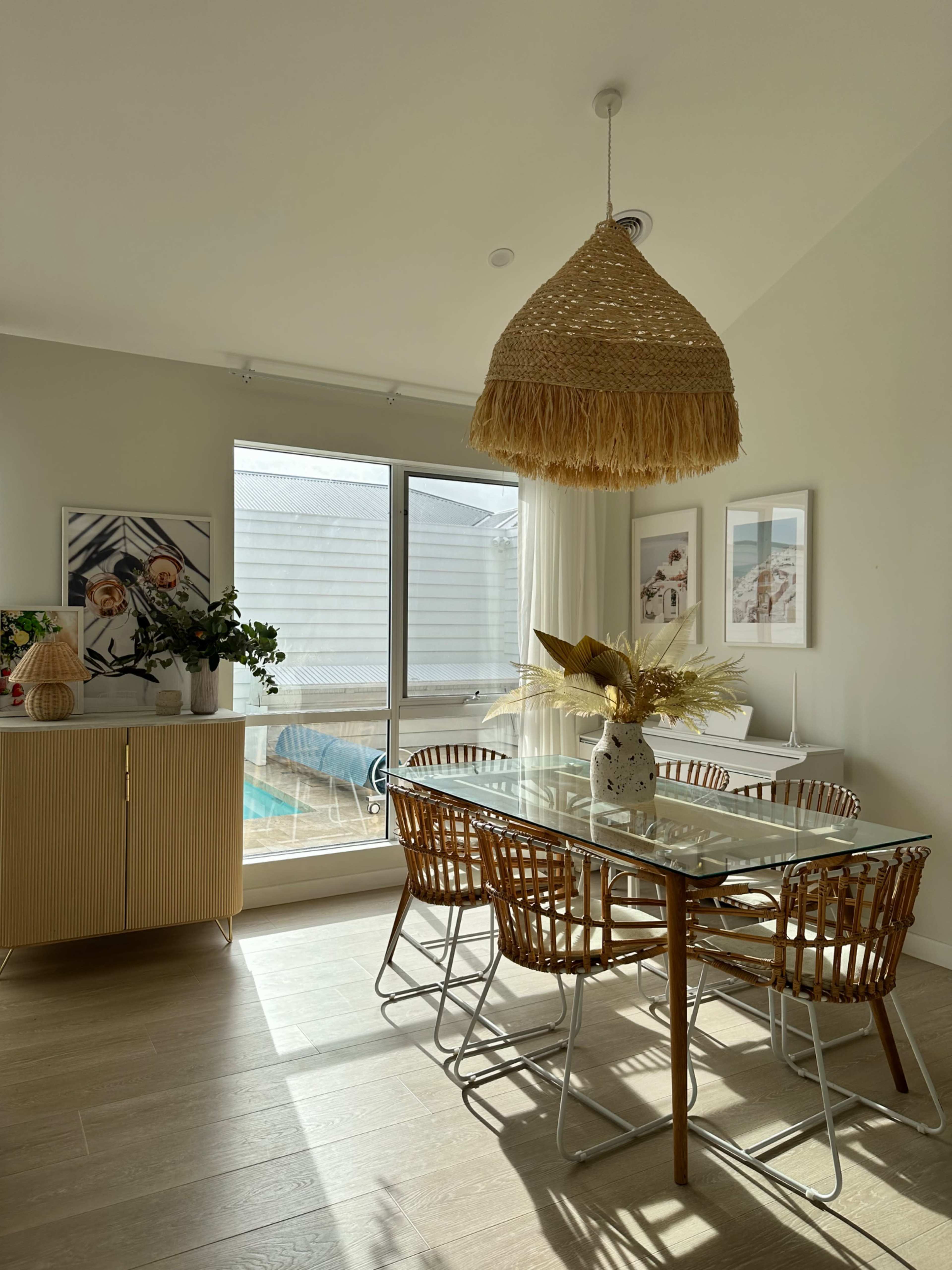 A dining area features a glass table surrounded by wicker chairs, a decorative cabinet, and large windows letting in natural light.