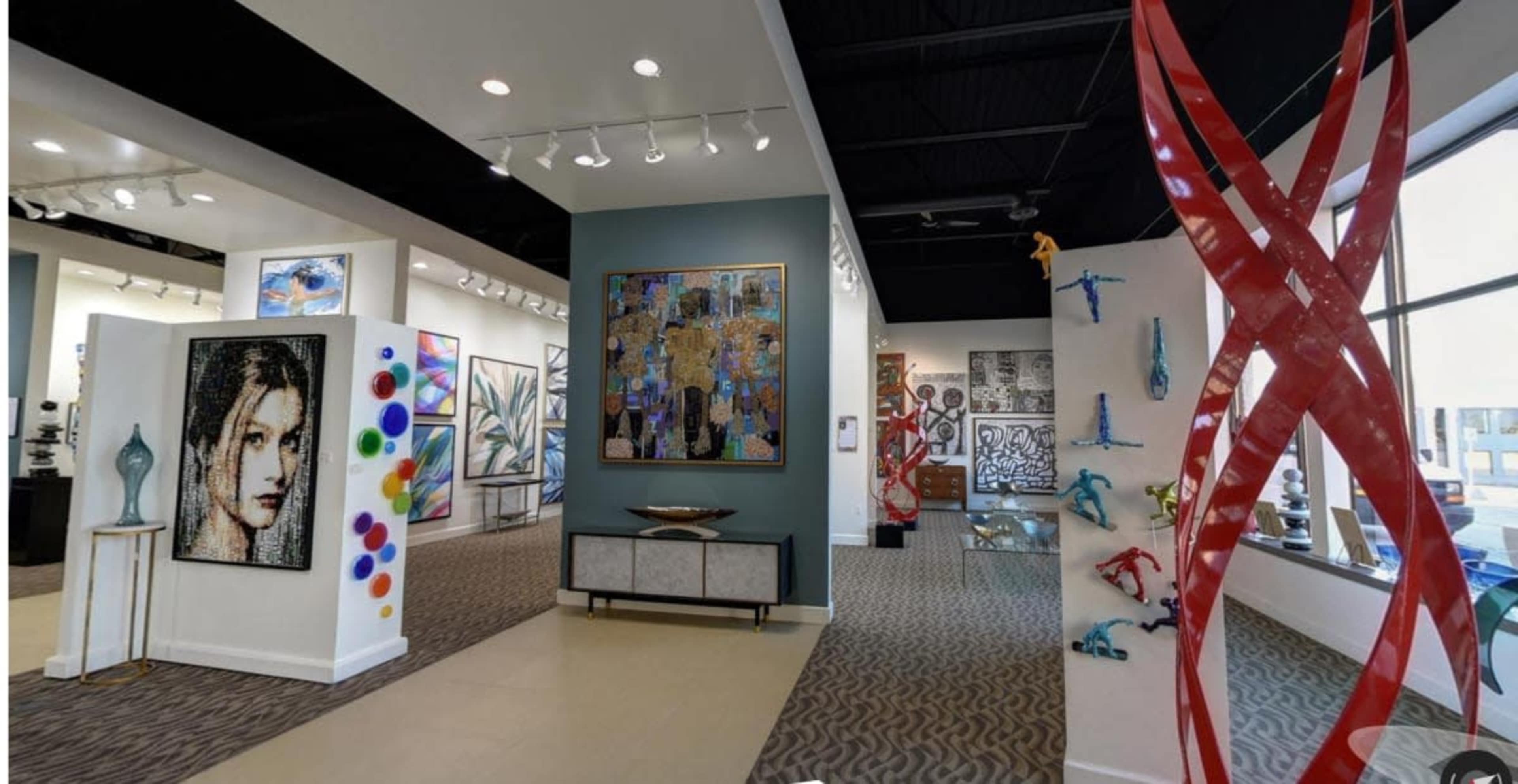 The image shows an art gallery interior with various artworks displayed on the walls, including paintings and sculptures, in a well-lit environment.