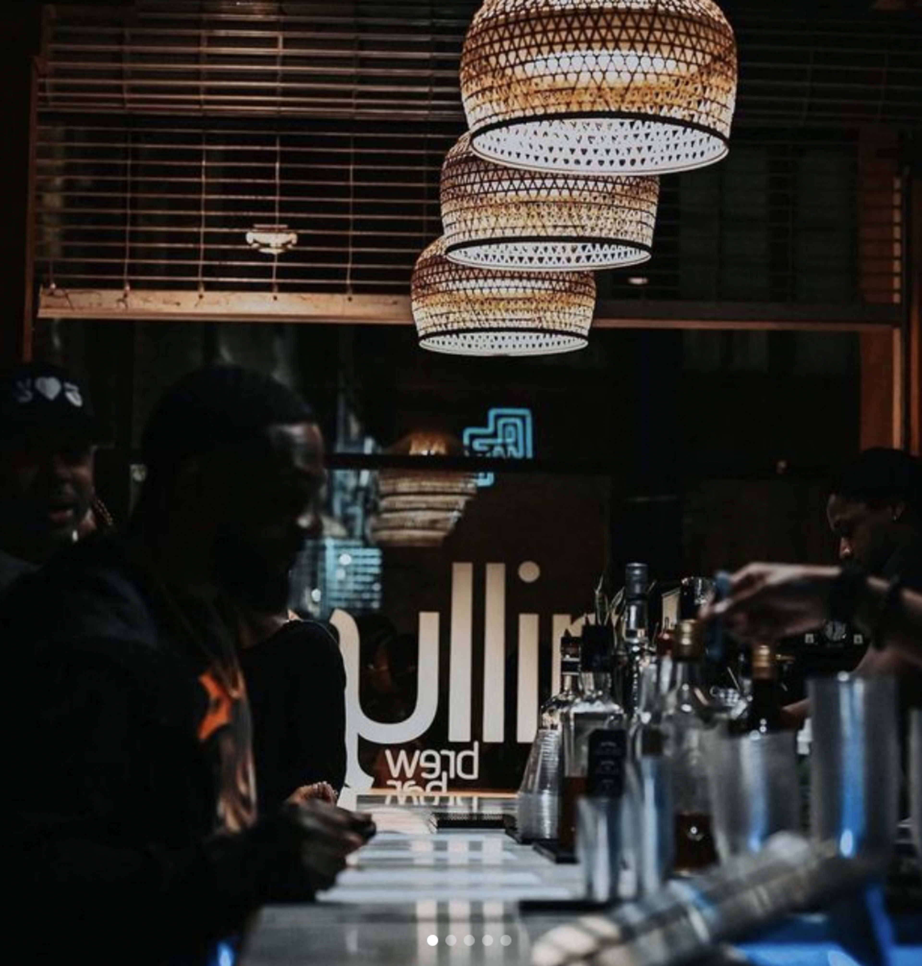 A nighttime scene shows patrons seated at a bar under woven pendant lights, with various bottles displayed on the counter.