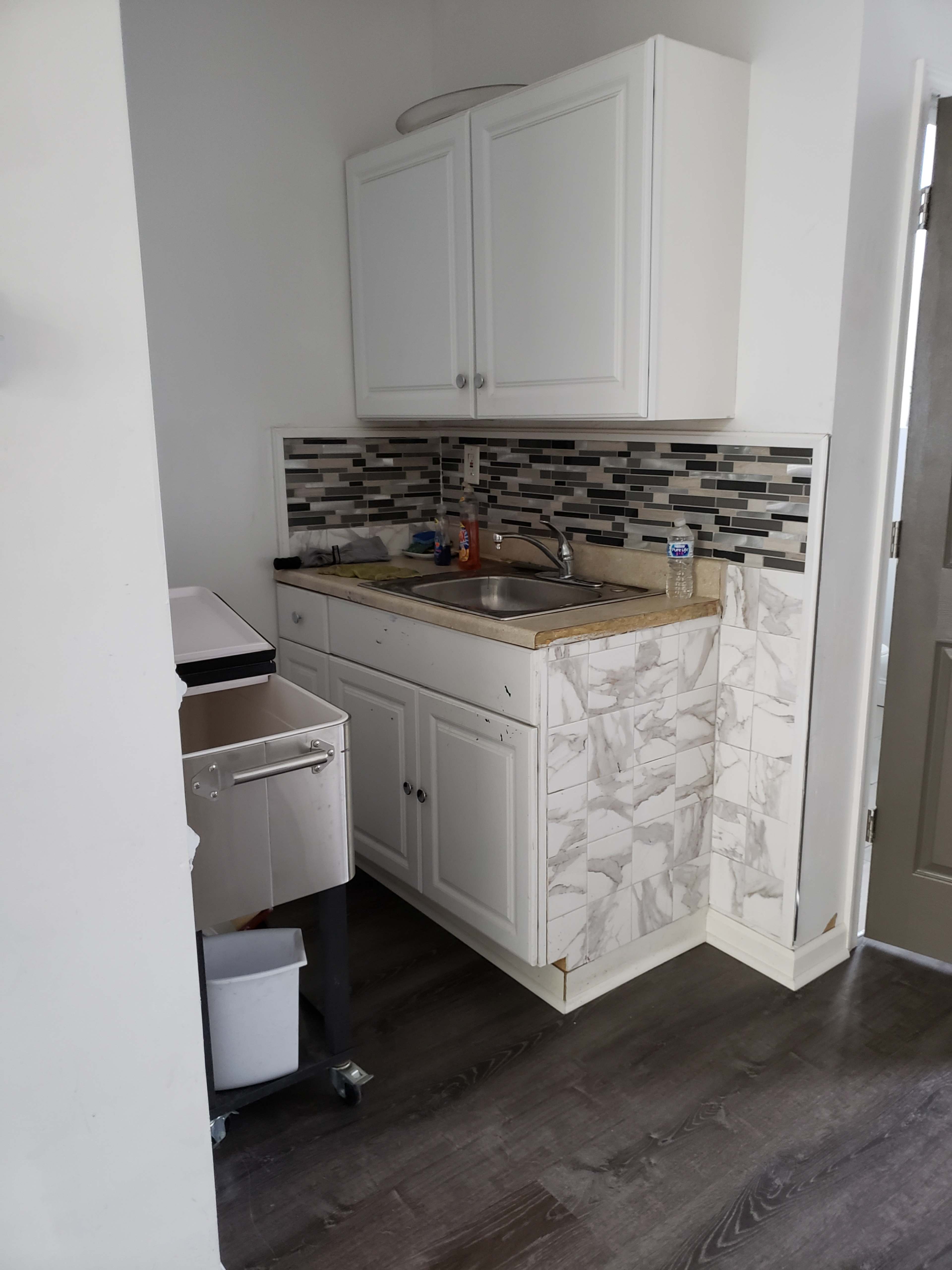 A small kitchenette features white cabinets, a sink, and a countertop with a marble design, alongside a mosaic backsplash.