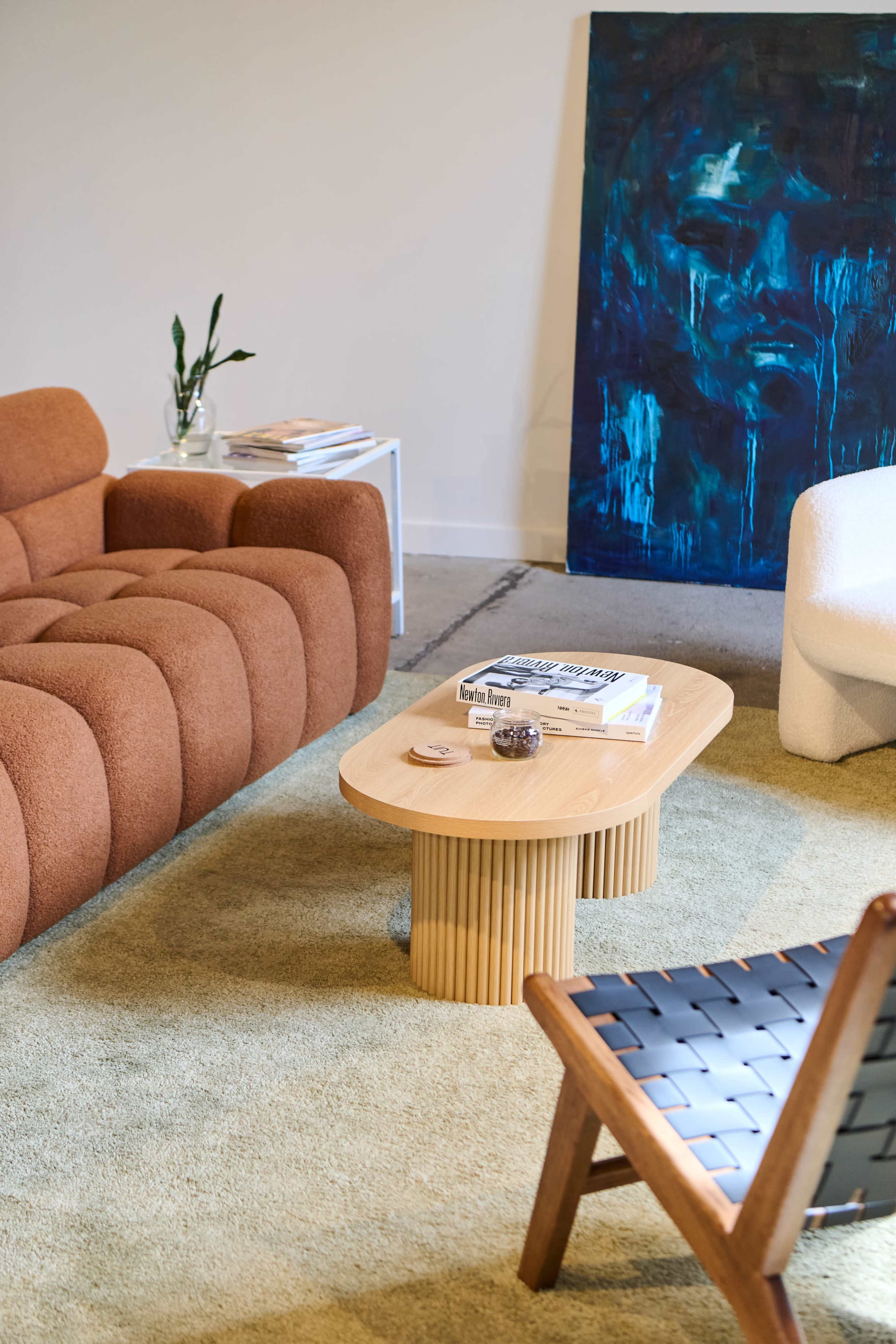 The image shows a modern living room featuring a brown curved sofa, a wooden coffee table with a fluted design, and a white sculptural chair, along with a blue artwork on the wall.