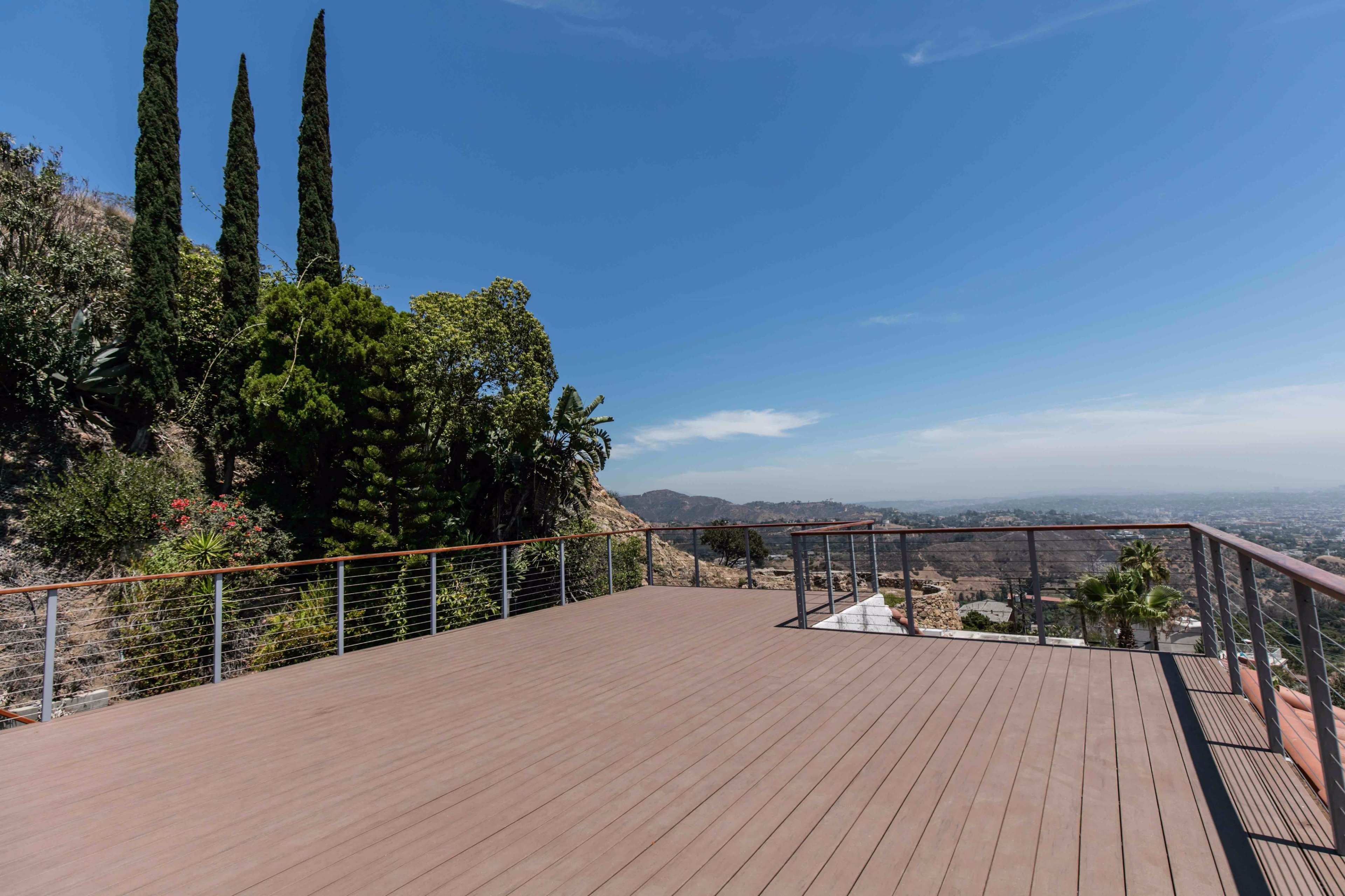 A spacious wooden deck overlooks a valley and is surrounded by various plants and trees under a clear blue sky.