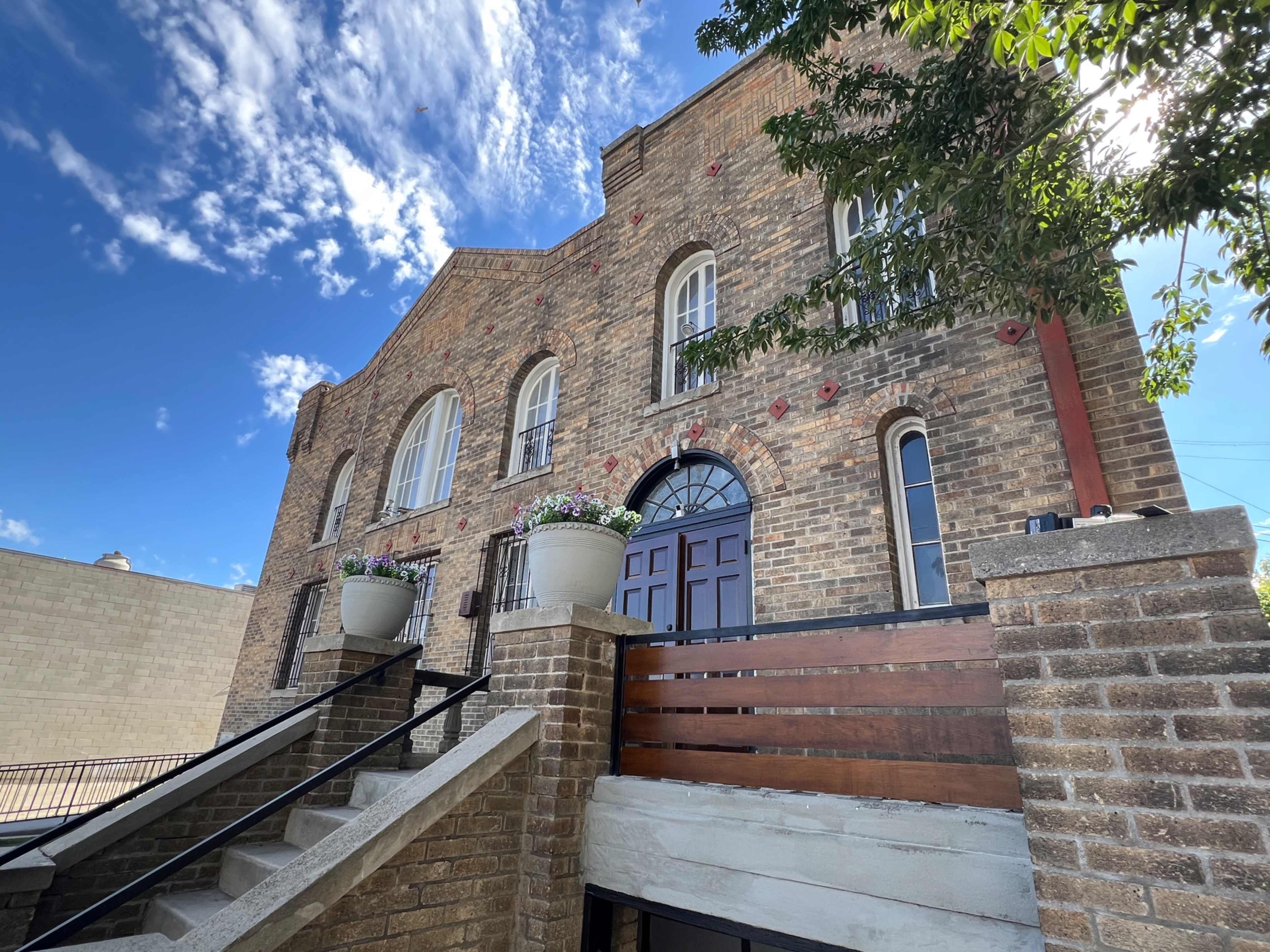 The image shows a brick building with large windows and decorative planters, situated at the top of a staircase under a blue sky with scattered clouds.