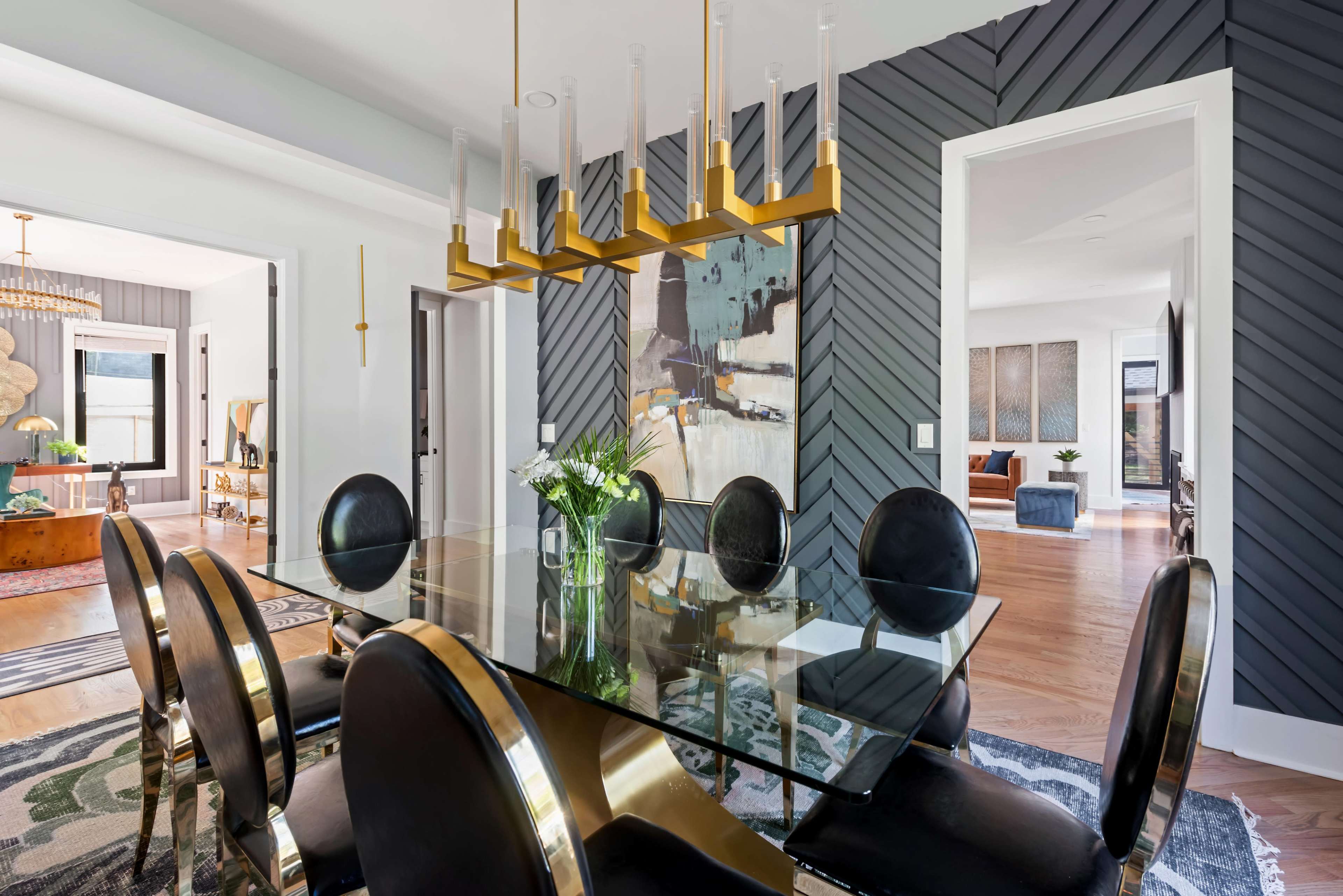 A modern dining room features a glass table surrounded by black upholstered chairs and a gold chandelier, with a patterned wall in the background.