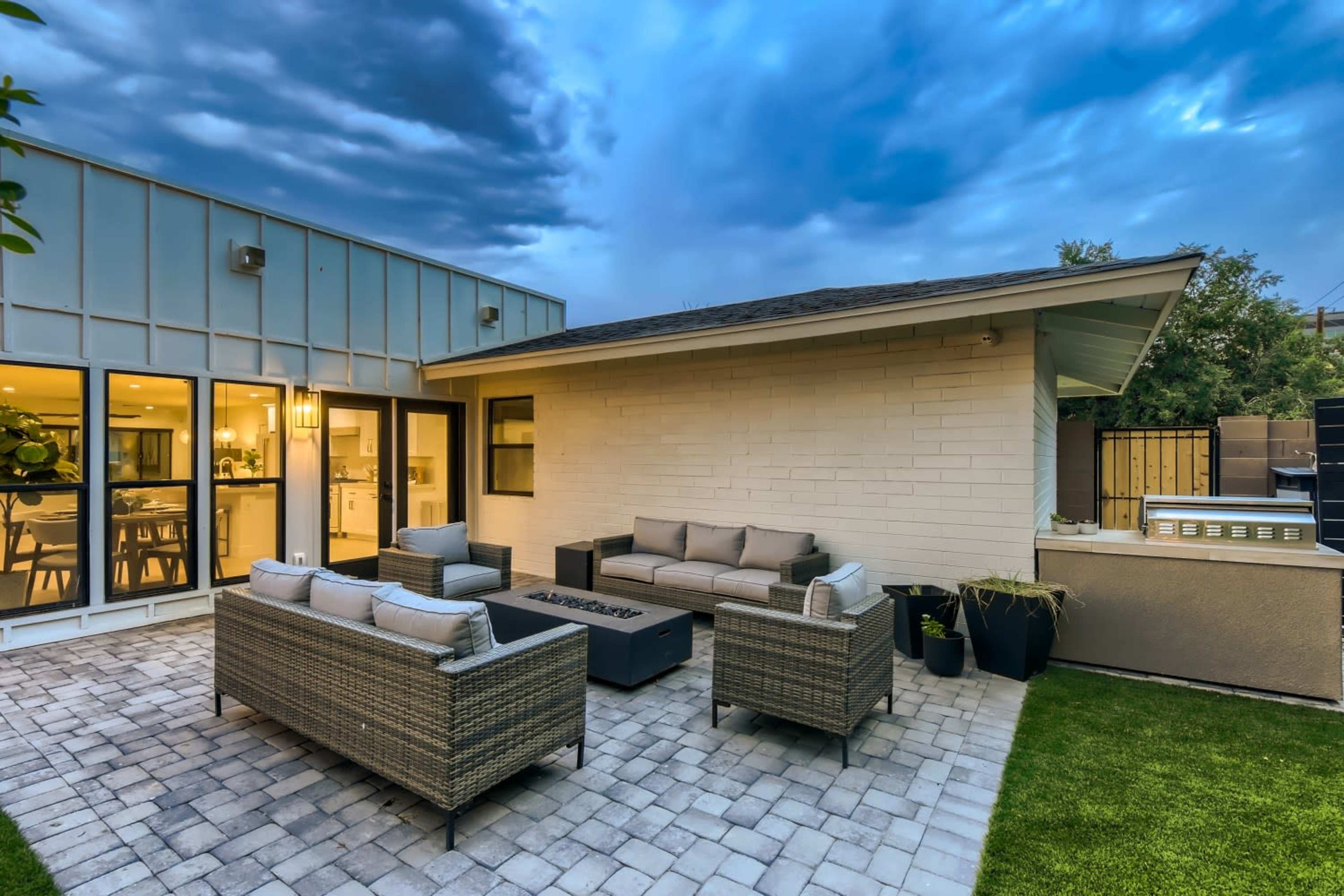A modern outdoor patio features wicker furniture arranged around a fire pit, with a view of a house and cloudy sky in the background.