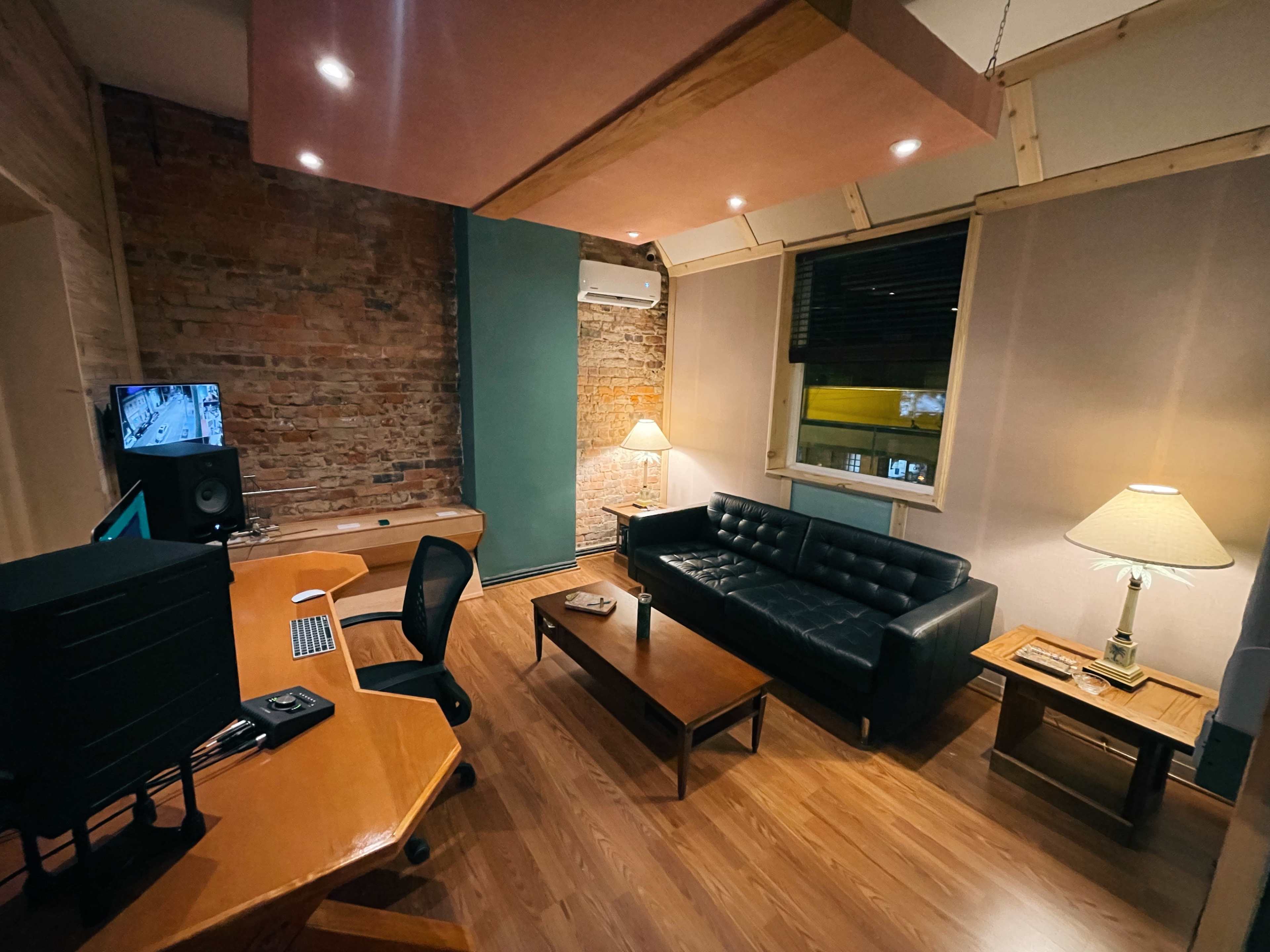 A cozy room with a black couch, a wooden coffee table, an office desk with a computer, and two lamps, featuring exposed brick walls and a large window.