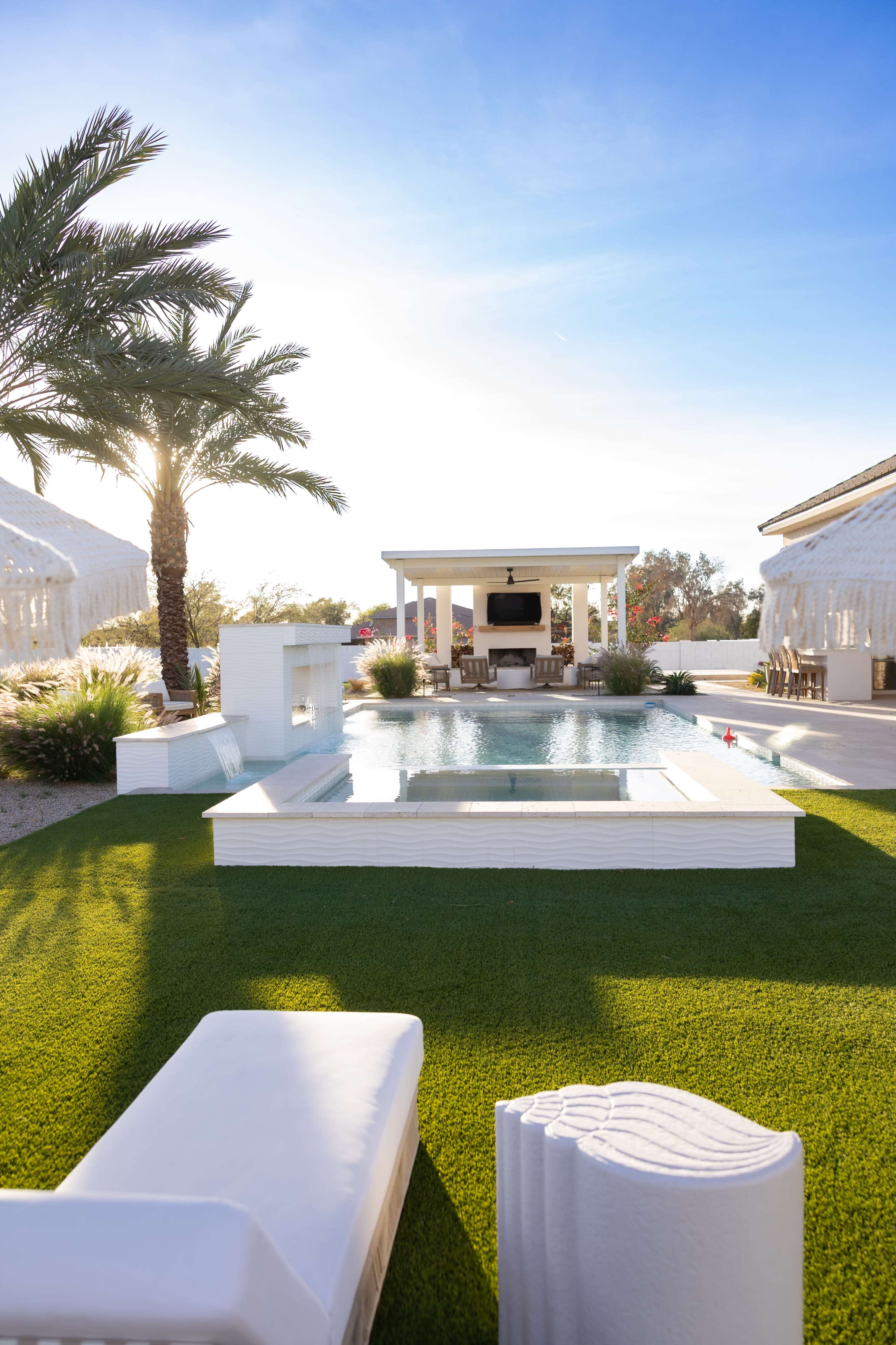 The image shows a modern outdoor space featuring a swimming pool, a well-manicured lawn, and a shaded seating area with palm trees in the background.