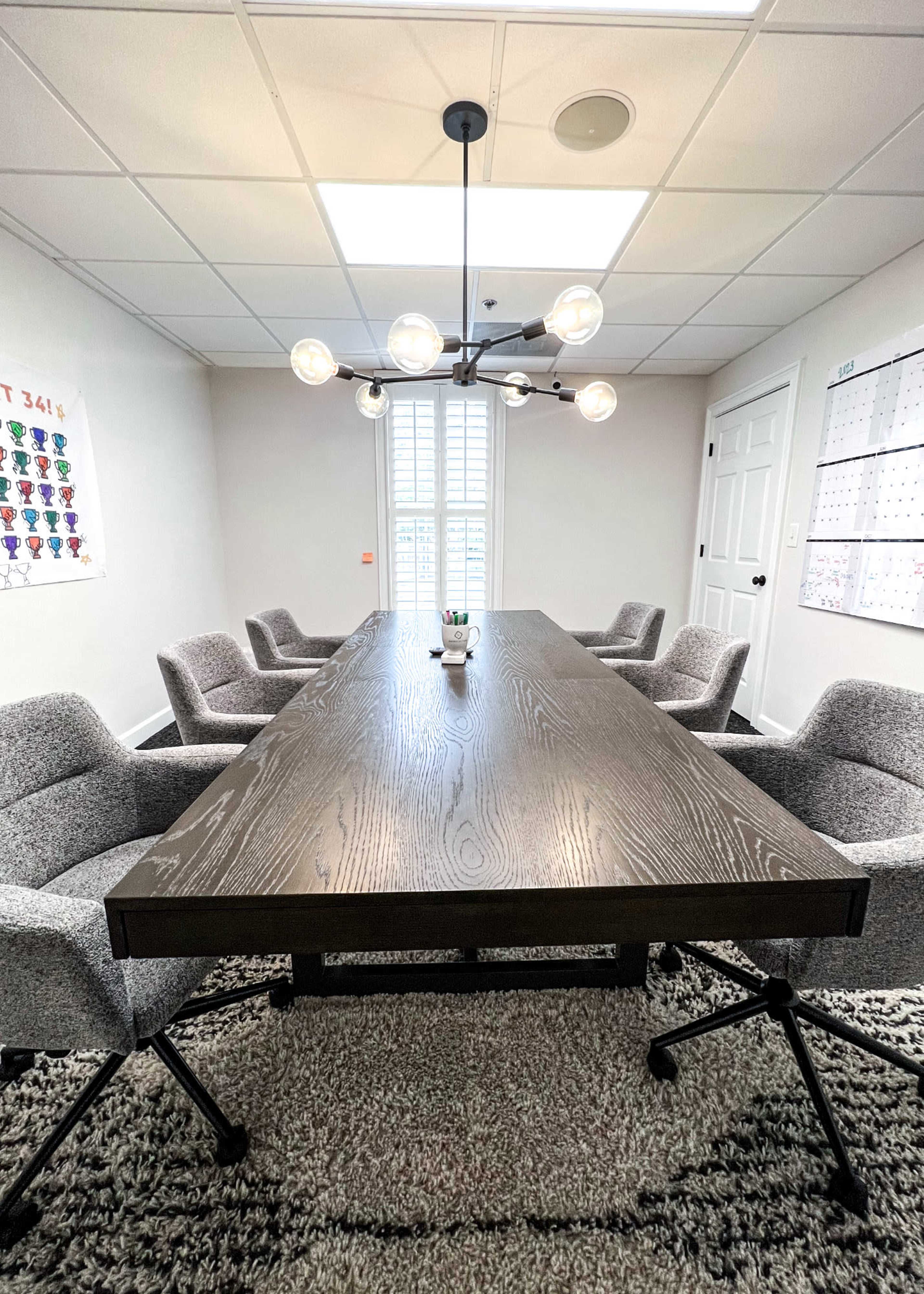 A large wooden conference table is surrounded by five gray chairs in a well-lit meeting room with a chandelier above.