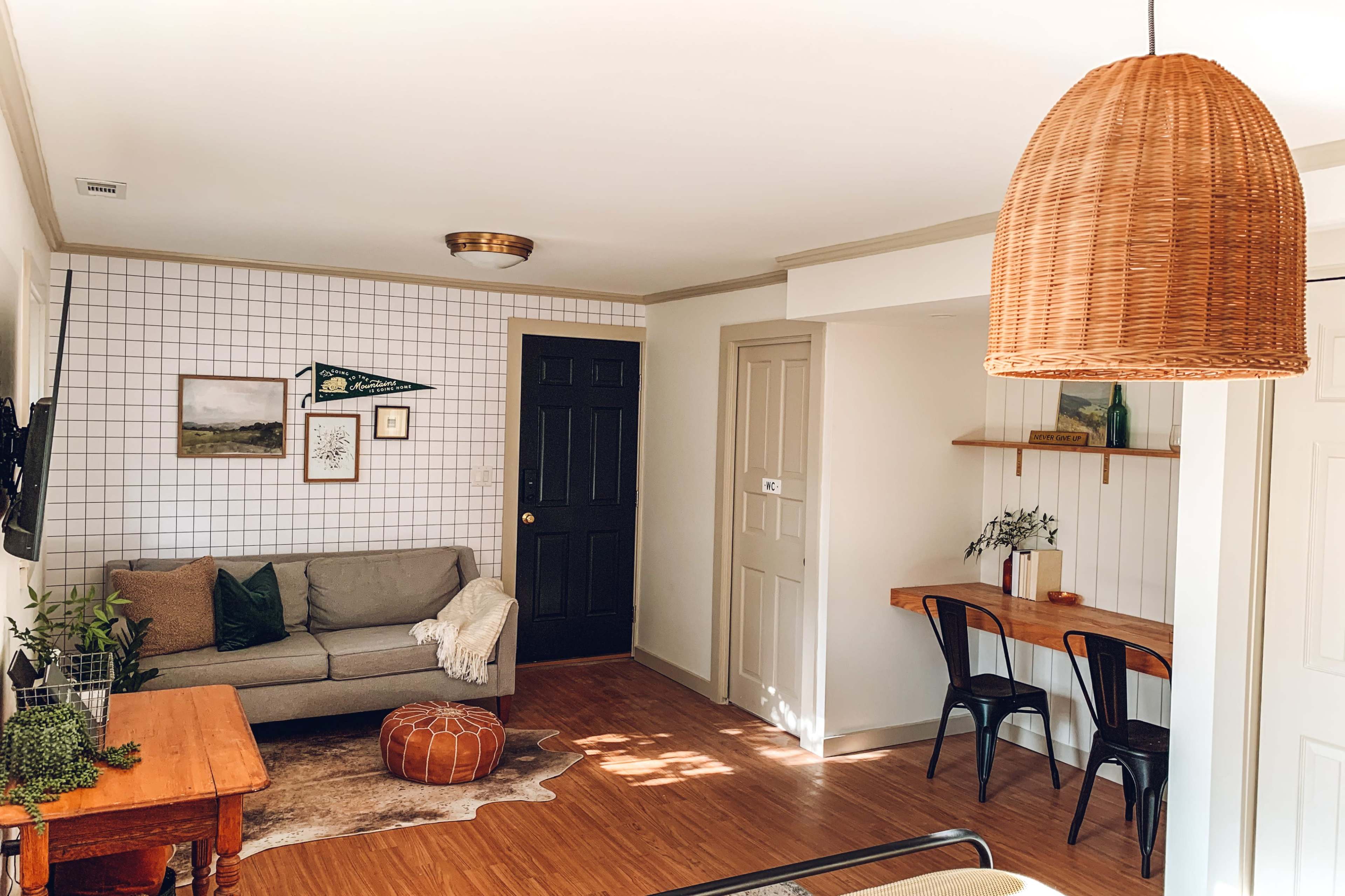 A living room features a gray sofa, a wooden table with black chairs, and a decorative pouf, all set against a checkered wall and warm wooden floors.