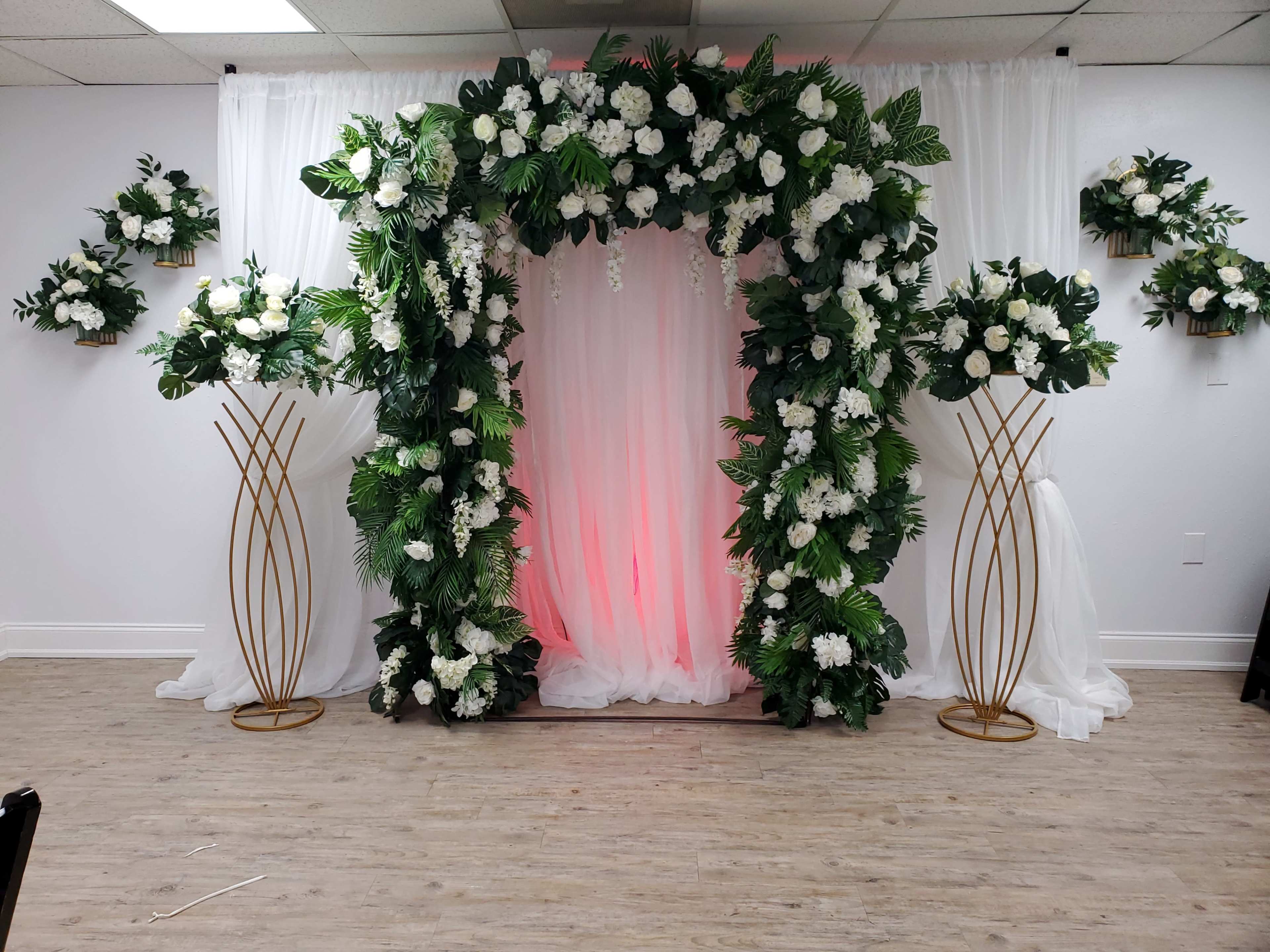 The image shows a decorated archway with white flowers and greenery, flanked by two gold vase stands, set against a draped white backdrop.