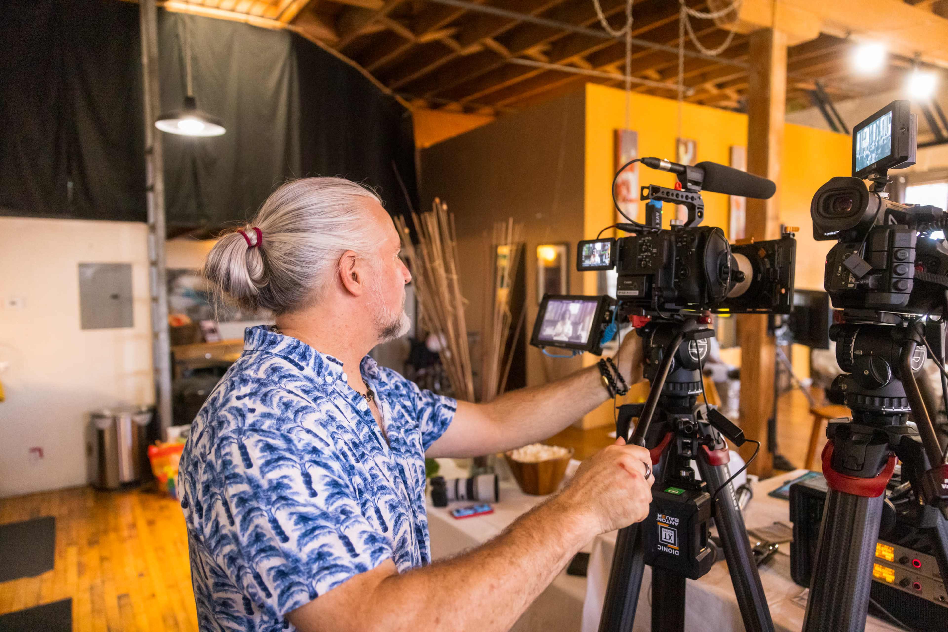A man with long hair tied back adjusts camera equipment in a well-lit interior space.