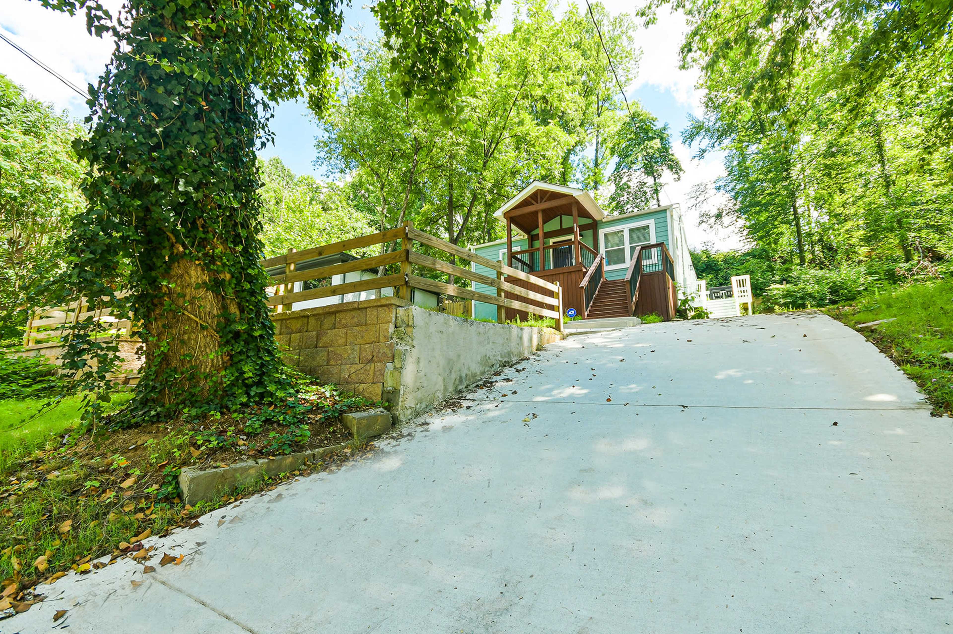 A house with a front porch is situated at the top of a sloped driveway surrounded by trees and greenery.